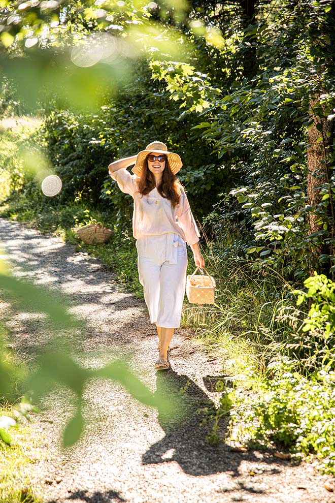 Woman in a sun hat and light linen outfit walking along a green forest path