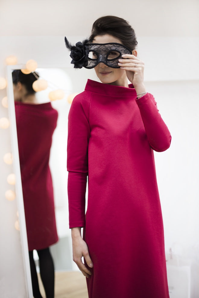 Woman in a magenta long-sleeve dress holding a masquerade mask by a mirror
