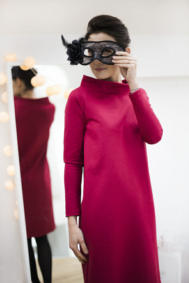 Kvinde i magenta langærmet kjole med en maskerademaske ved et spejl