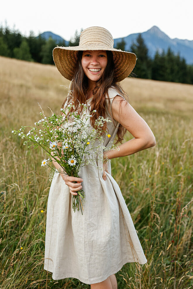 Smiling woman in a natural linen dress and straw hat holding wildflowers in a meadow