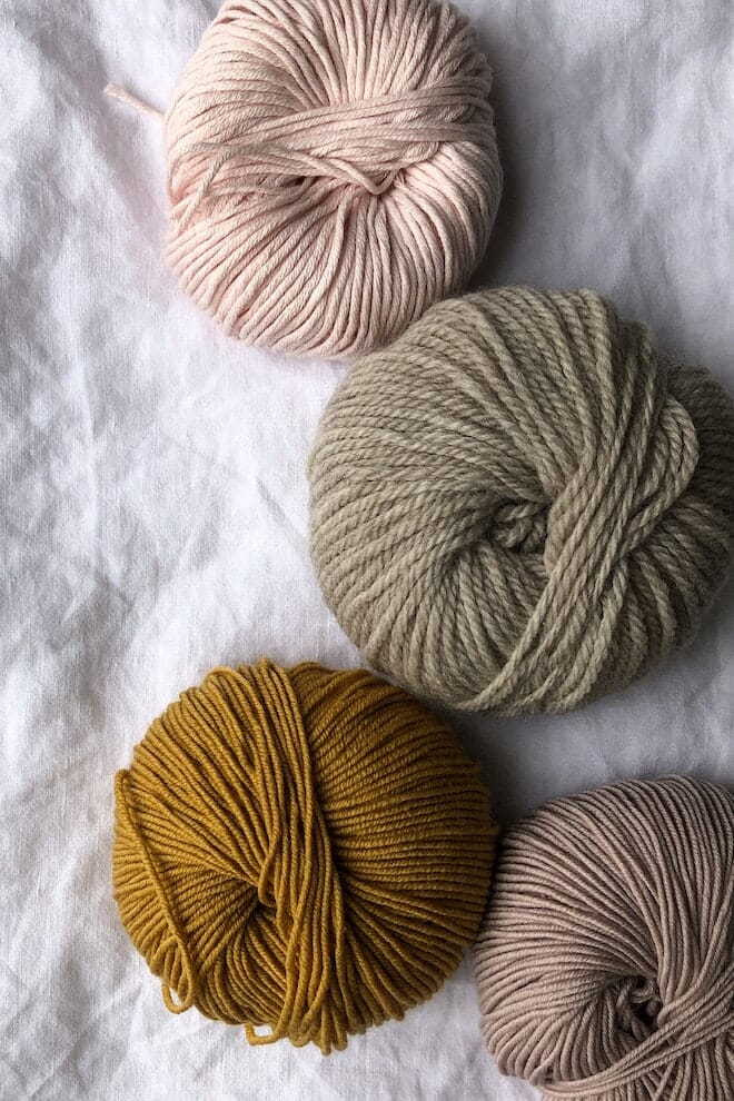 Skeins of wool yarn in cream, sage, mustard and taupe on white linen