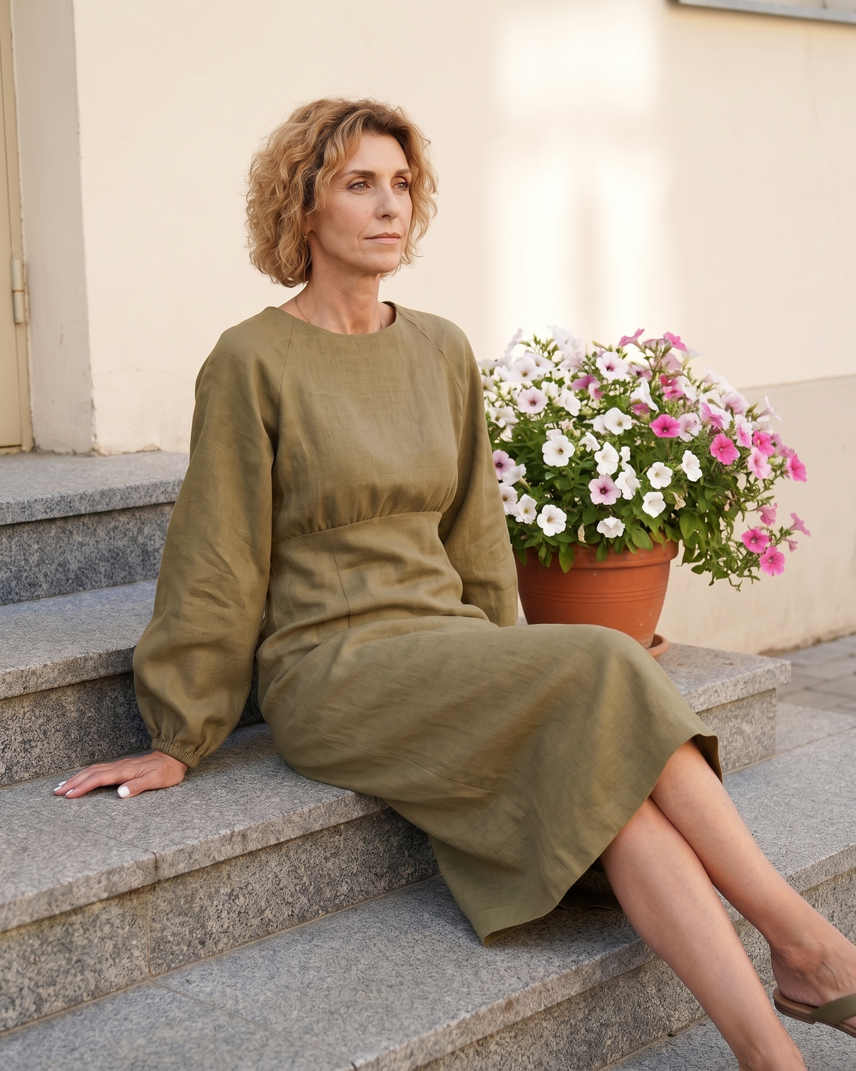 A woman sitting on stone steps next to potted flowers, wearing an olive green long-sleeve linen midi dress. #Color_Olive green