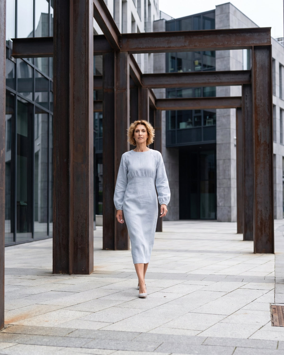 A woman walking on a city street wearing a light blue long-sleeve linen midi dress with a gathered waist, full length front view. #Color_Light blue