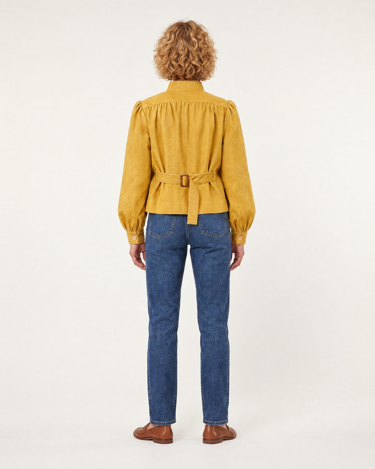 A woman stands with her back to the camera against a plain light studio background, wearing a mustard-ochre linen jacket with a stand collar, gathered shoulders, and a tie belt with buckle at the back, paired with blue jeans and brown loafers. #Color_Ochre