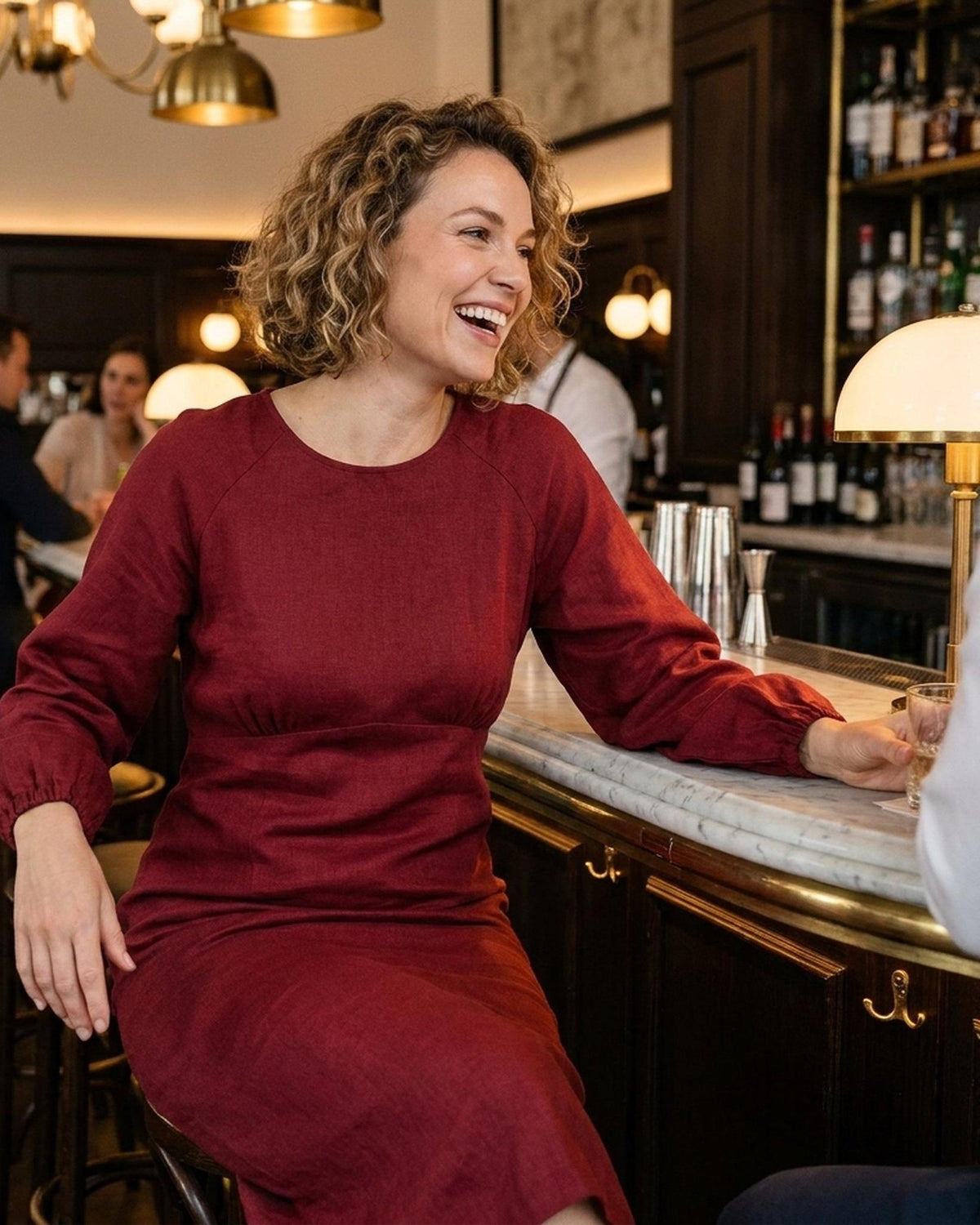 A woman sitting at a bar counter, smiling, wearing a long-sleeve burgundy linen midi dress with a gathered waist. #Color_Burgundy