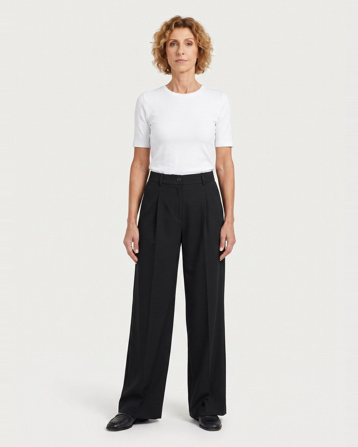 A woman stands against a light gray studio background wearing a white short-sleeve crewneck T-shirt and black wide-leg trousers. #Color_Black