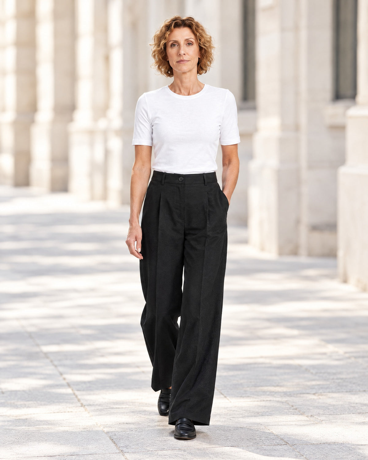 A woman walks along a sunlit colonnade wearing a white short-sleeve crewneck top and black wide-leg trousers with black loafers. #Color_Black