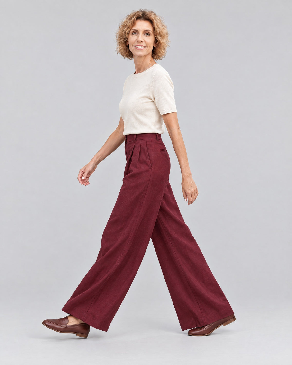 A woman walks mid-stride in profile against a light gray studio background, wearing a cream short-sleeve top, burgundy wide-leg trousers and brown loafers. #Color_Burgundy