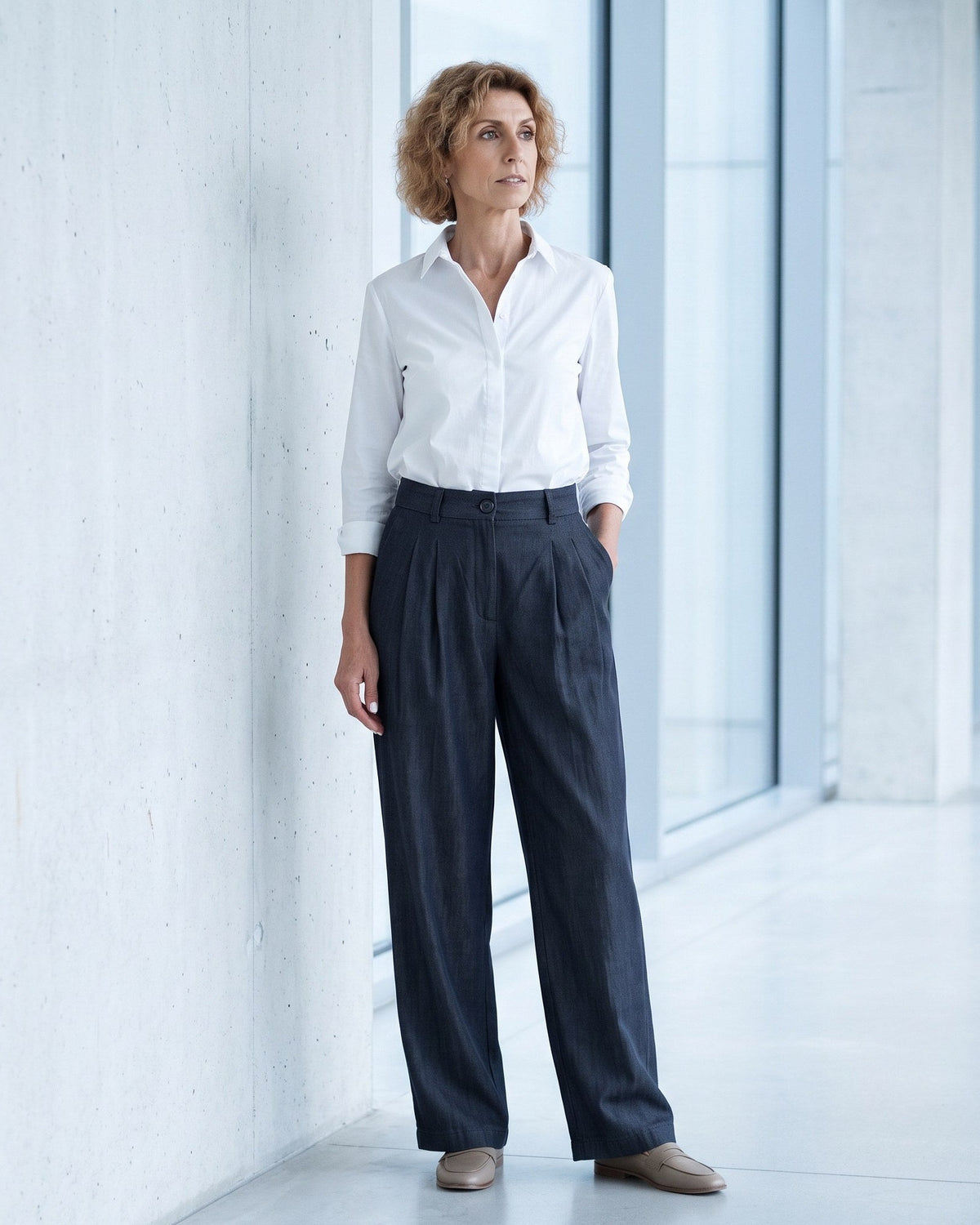A woman stands in a bright, modern indoor corridor wearing a white button-up shirt tucked into dark navy wide-leg trousers, paired with tan shoes. #Color_Deep blue