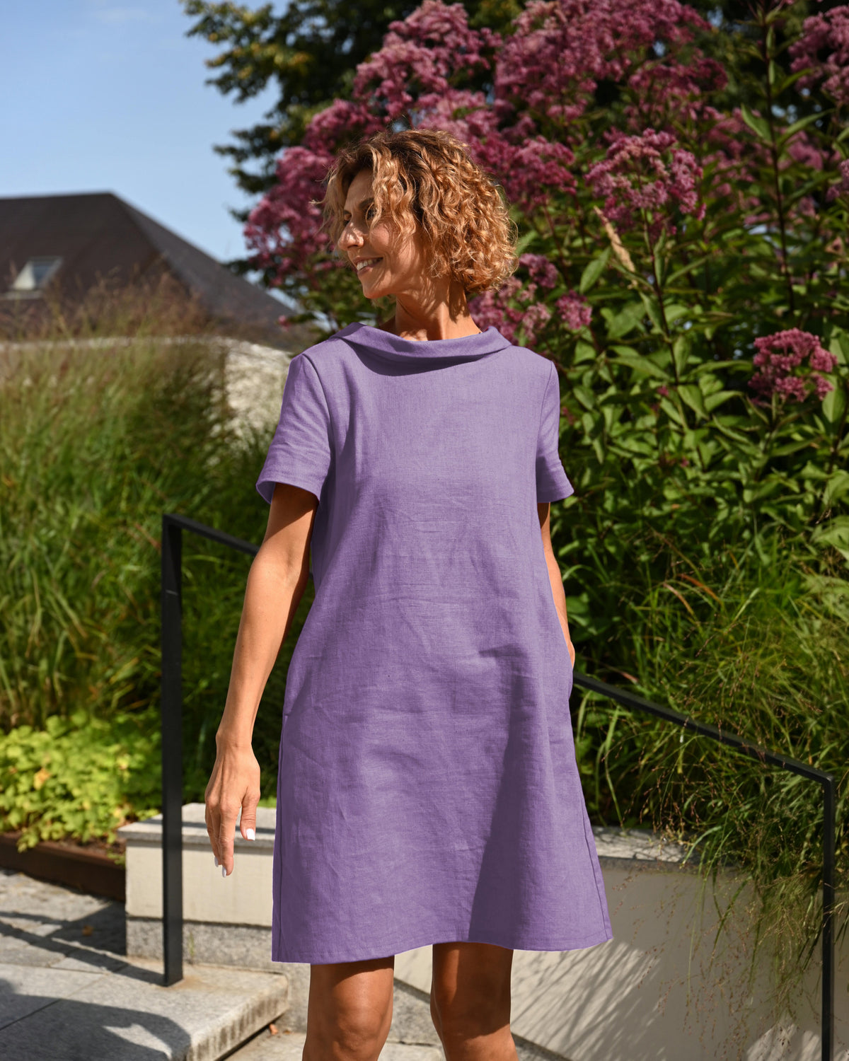 LeMuse STORY linen dress in purple – front view by flowering shrub #Color_Purple