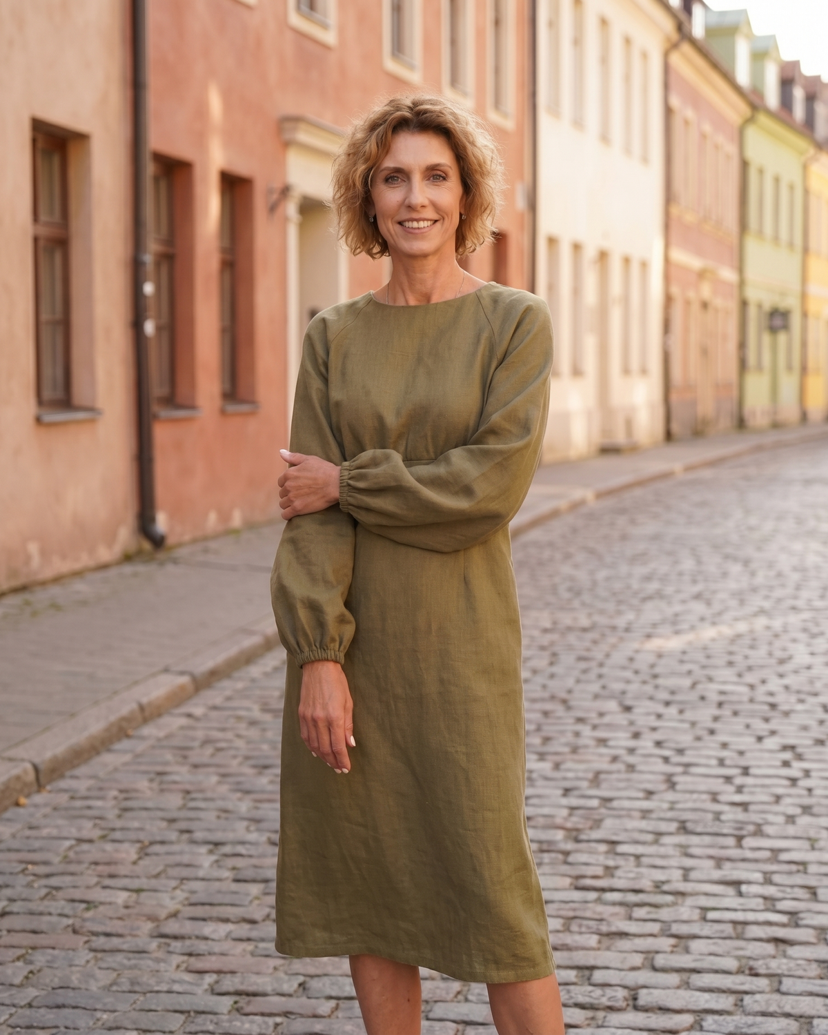 A woman standing on a cobblestone street, smiling, wearing an olive green long-sleeve linen midi dress with a gathered waist. #Color_Olive green