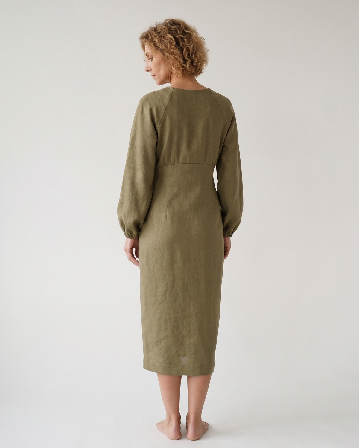 A woman viewed from the back wearing an olive green long-sleeve midi dress with a gathered waist, shown against a plain studio background. #Color_Olive green