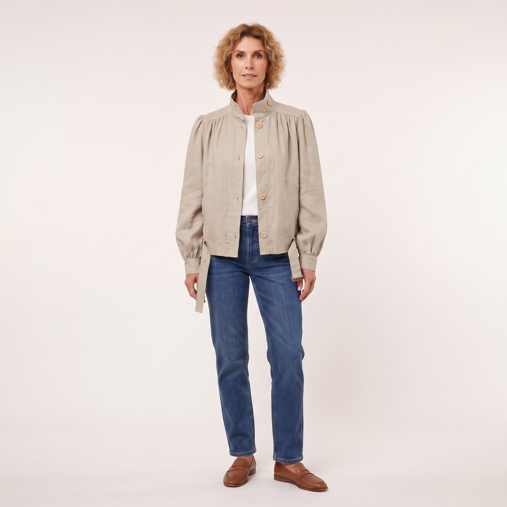 A woman modeling a light beige linen jacket with gathered shoulders and gold buttons, worn over a white top and blue jeans, against a plain off-white studio background. #Color_Beige