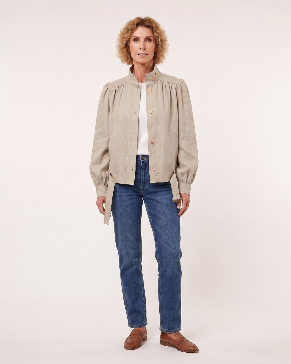 A woman modeling a light beige linen jacket with gathered shoulders and gold buttons, worn over a white top and blue jeans, against a plain off-white studio background. #Color_Beige