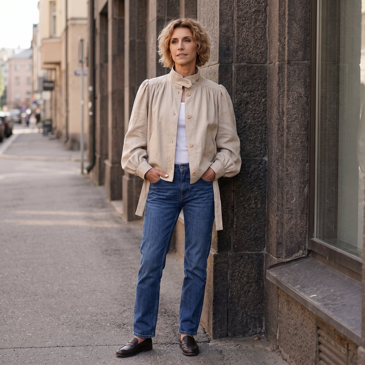 A woman stands on a city sidewalk leaning against a dark stone building, wearing a beige high-neck jacket over a white top with blue jeans and dark brown loafers. #Color_Beige