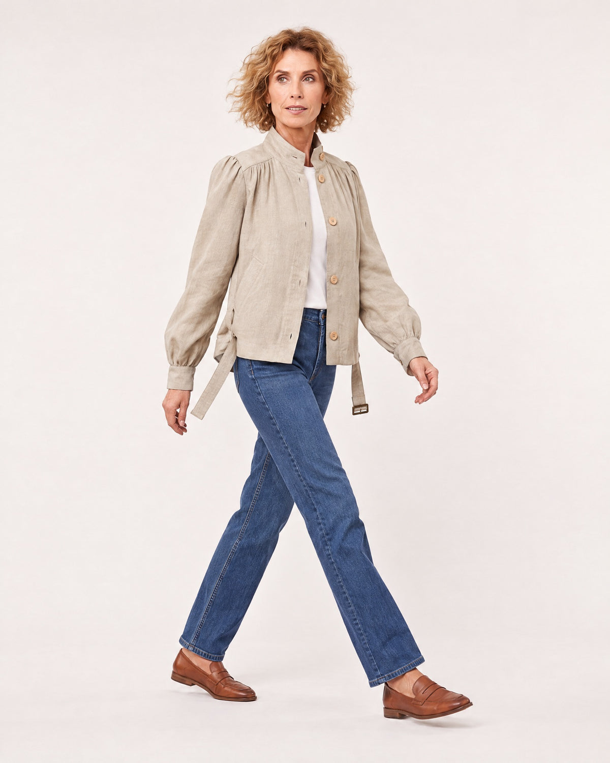 A woman walks in profile against a plain light studio background, wearing a light beige linen jacket with a stand collar and tie belt, paired with blue jeans and brown loafers. #Color_Beige