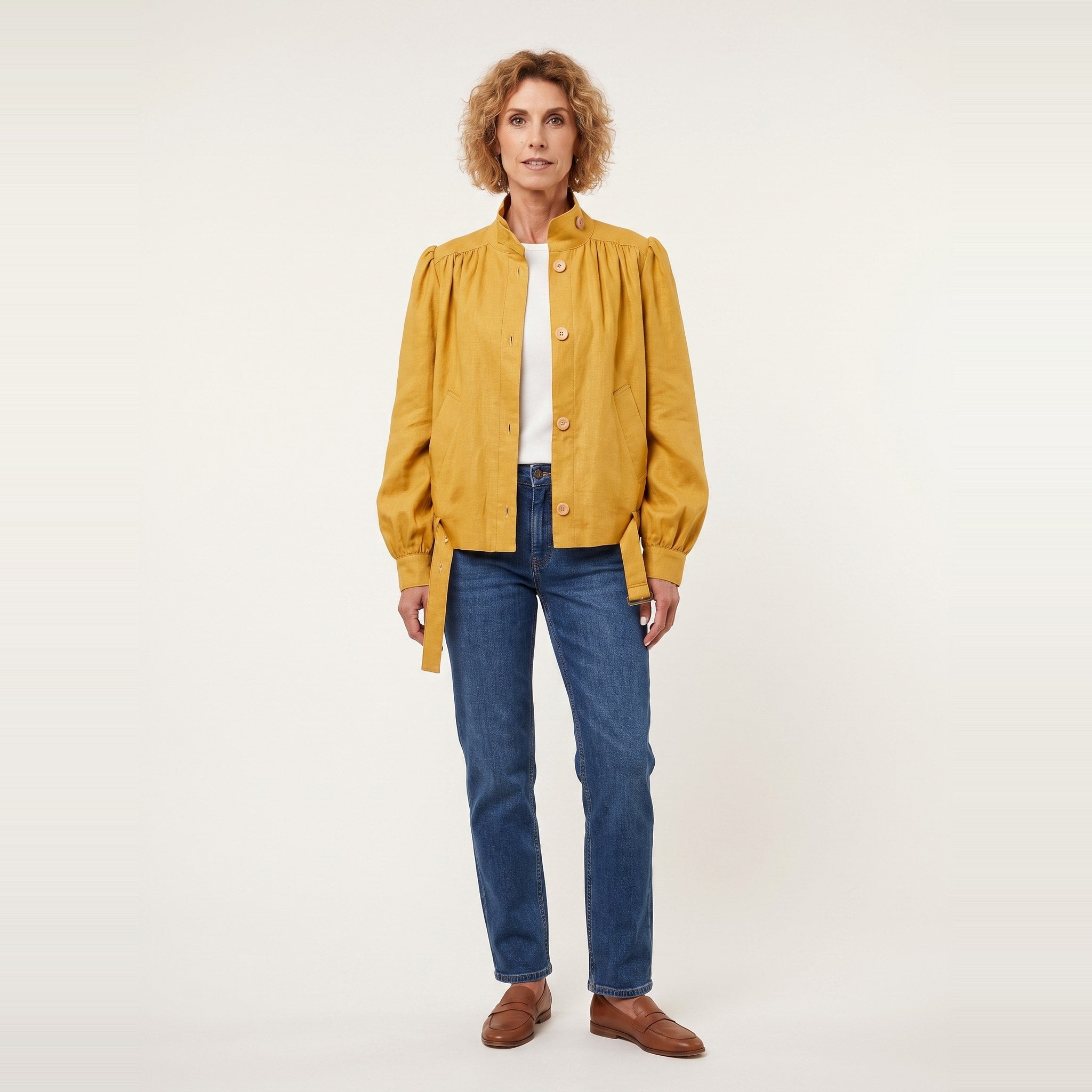 A woman stands against a plain light background wearing a mustard yellow button-front jacket with gathered sleeves, blue jeans, a white top, and brown shoes. #Color_Ochre