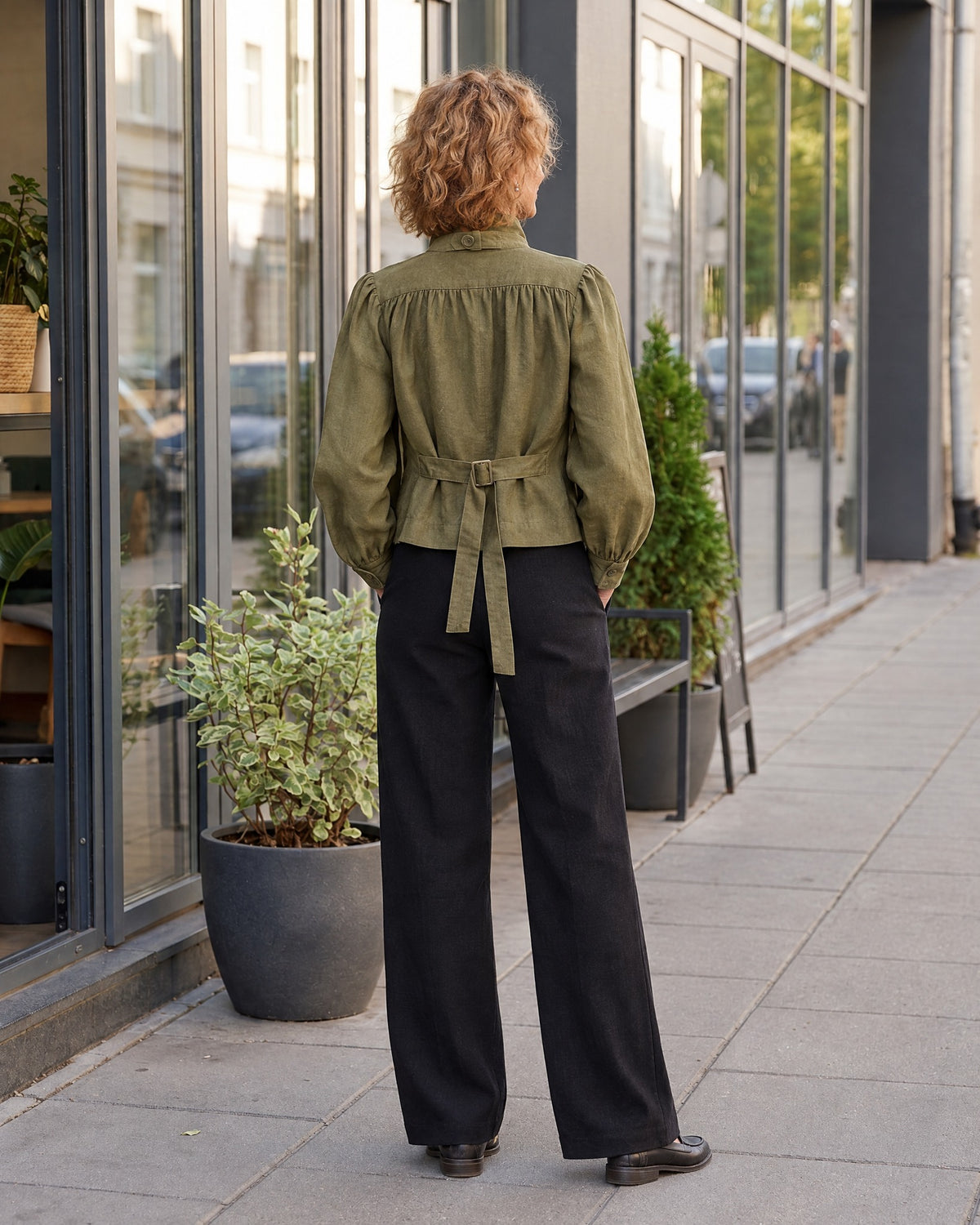 A woman stands with her back to the camera on a city sidewalk, wearing an olive green linen jacket with a stand collar, gathered shoulders, and a tie belt with buckle at the back, paired with black wide-leg trousers. #Color_Olive green