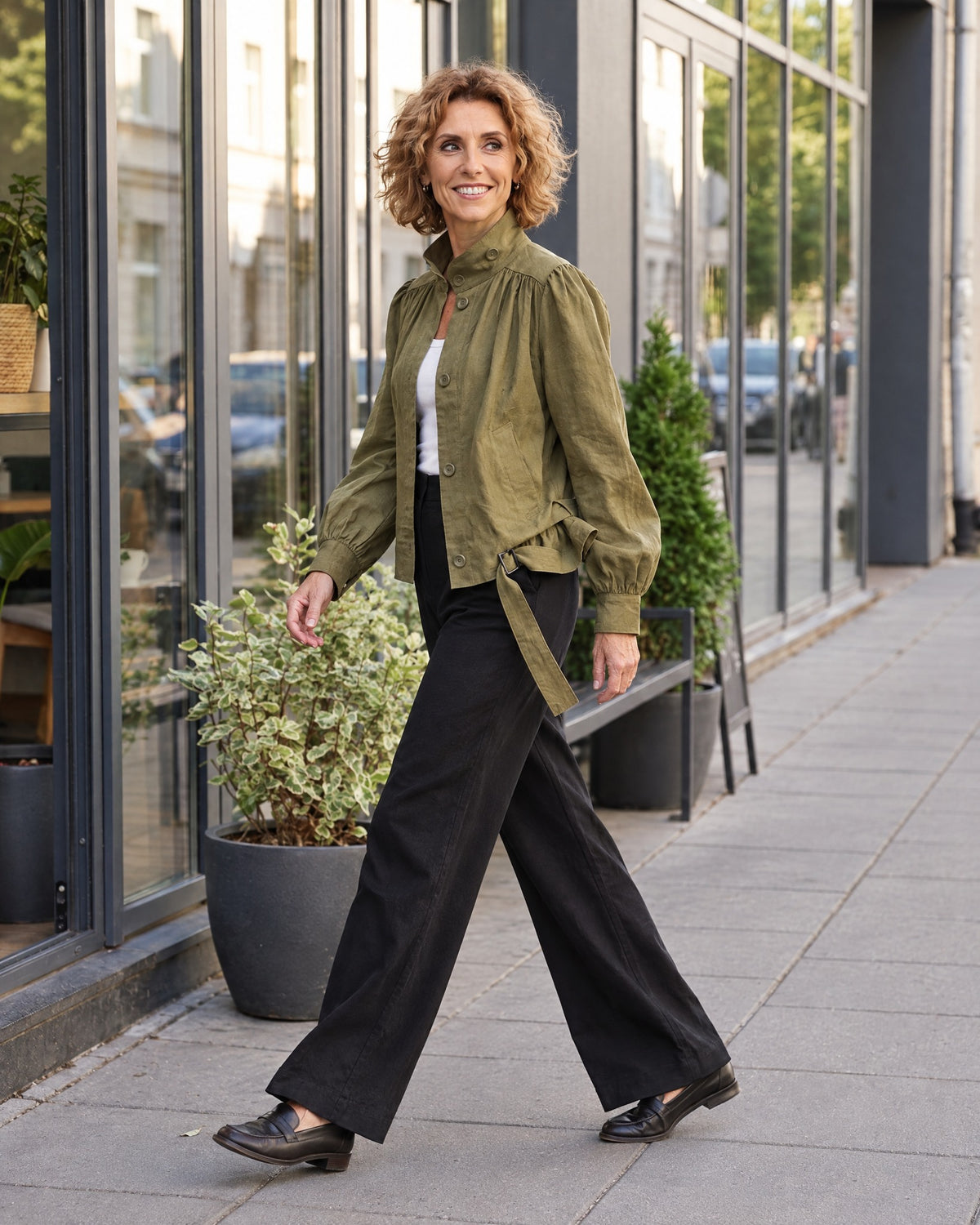 A woman walks along a city sidewalk in profile, wearing an olive green linen jacket with a stand collar and tie belt, paired with black wide-leg trousers and dark loafers.