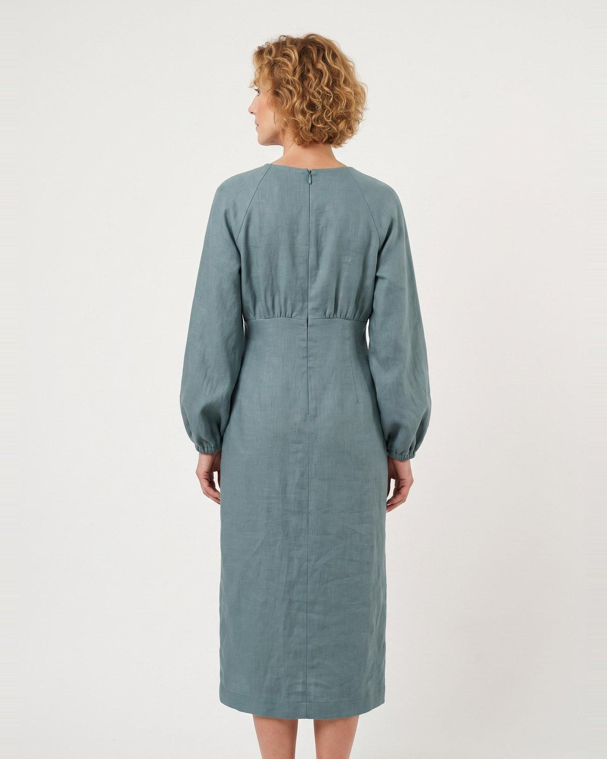 A woman viewed from the back wearing a long-sleeve midi dress in a muted dusty blue-green color, set against a plain off-white studio background. #Color_Teal