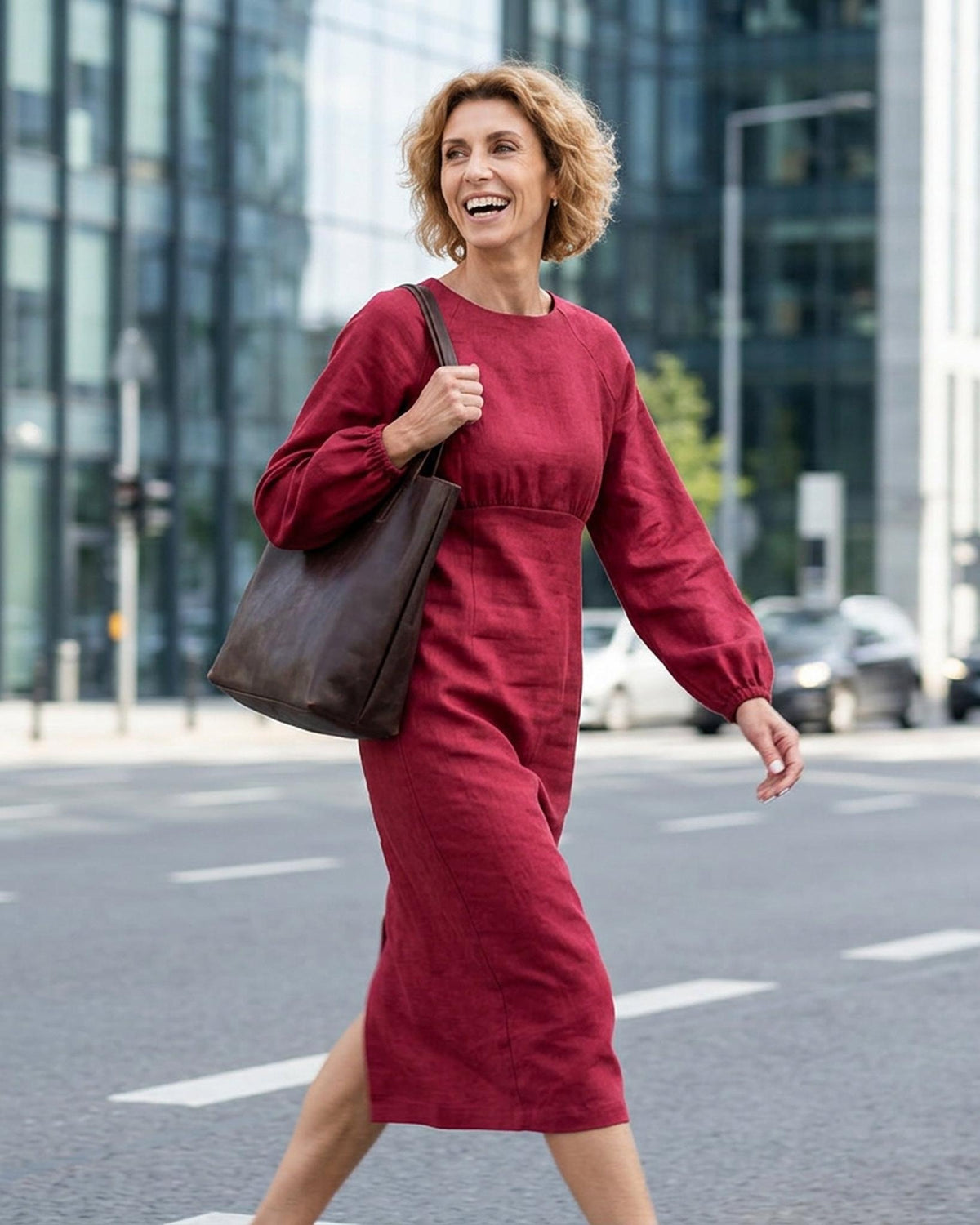 A woman walking across a city street wearing a long-sleeved, knee-length cerise red dress and carrying a large dark brown leather tote bag. #Color_Cerise