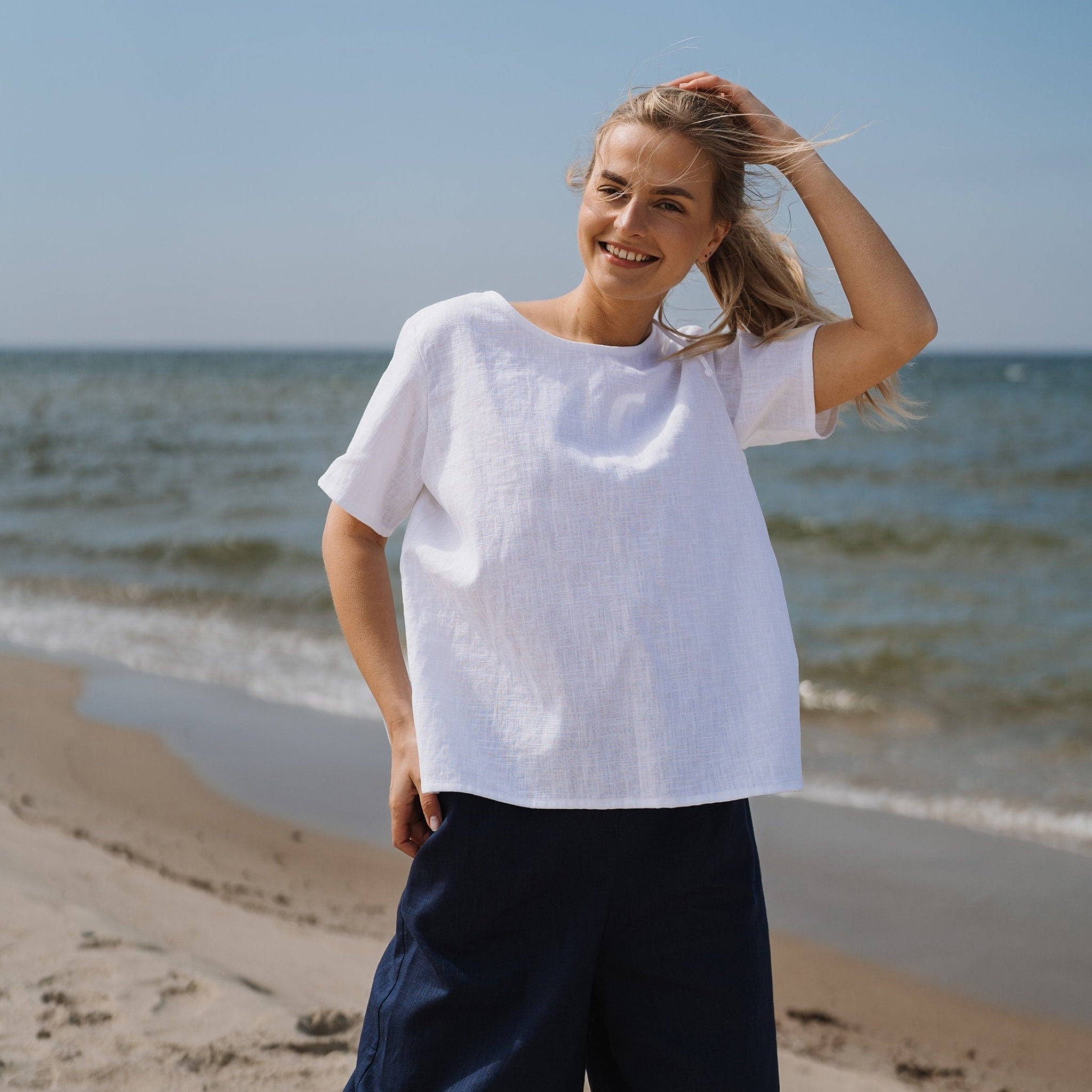 A woman stands on a sandy beach wearing a loose white short-sleeve top and dark navy pants, with the ocean and blue sky behind her.