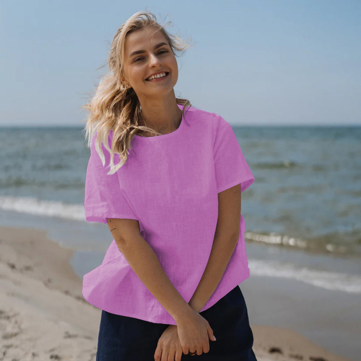 A woman with blonde hair stands on a sandy beach wearing a bright pink short-sleeve linen top and dark navy pants, with the ocean and blue sky in the background.