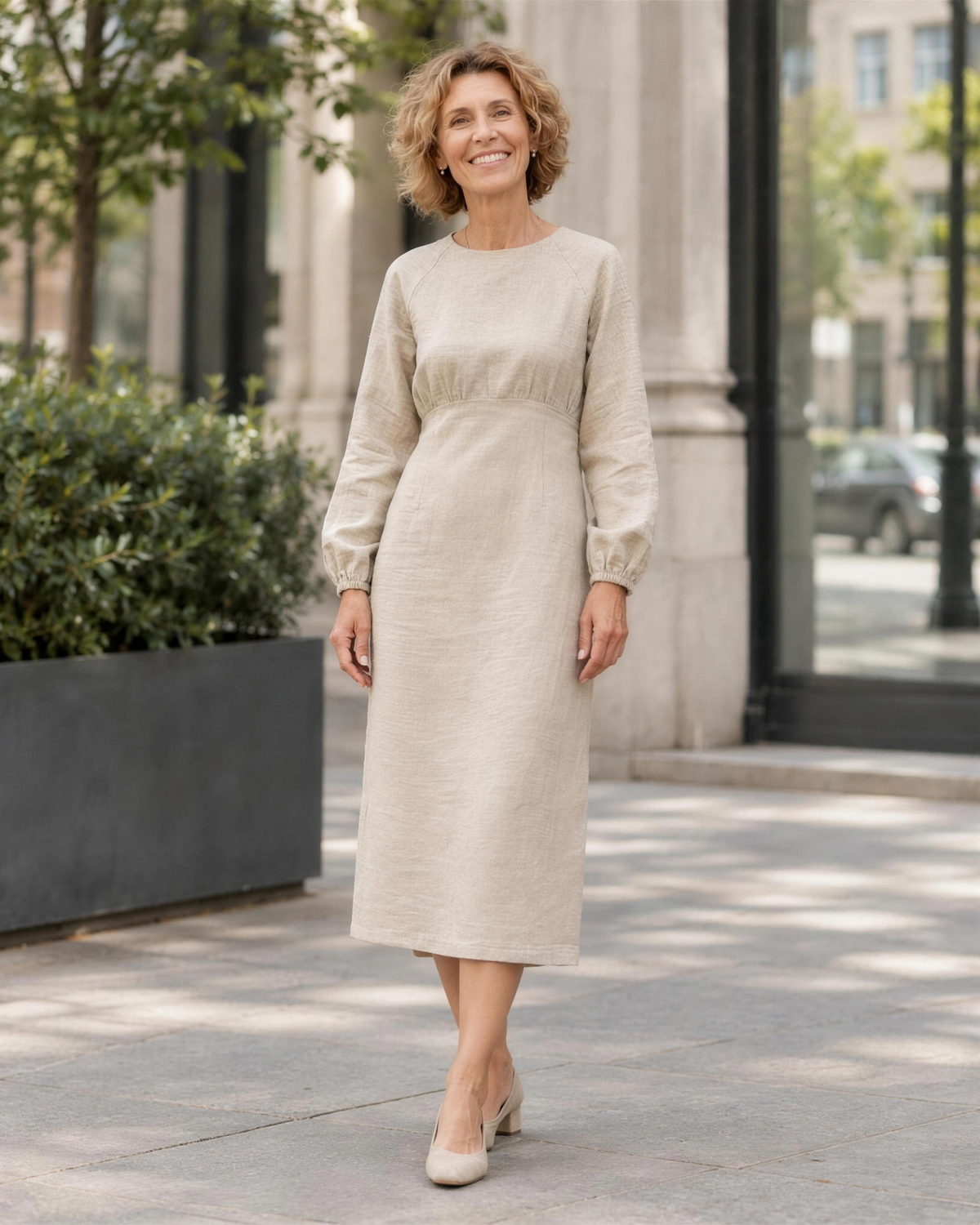 A woman walking on a city street, smiling, wearing a light beige long-sleeve linen midi dress with a gathered waist. #Color_Beige