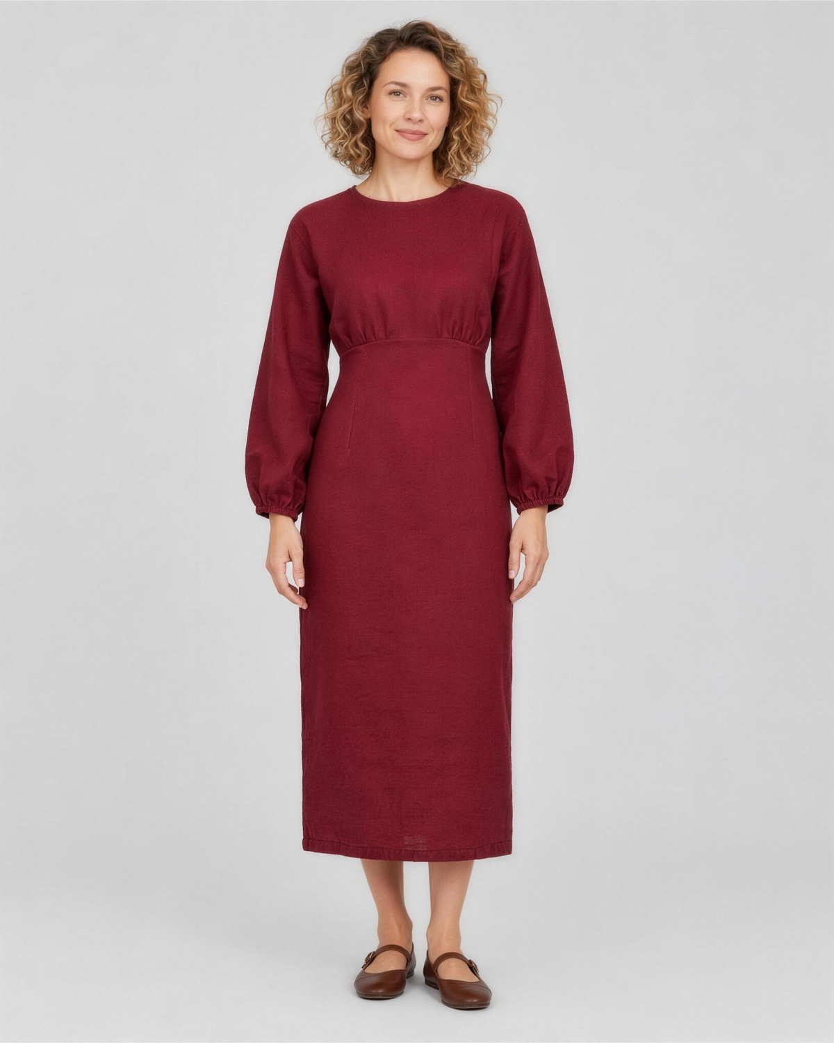 A woman standing in a studio, facing camera, full length, wearing a deep burgundy linen midi dress with a gathered waist and elastic cuffs. #Color_Burgundy