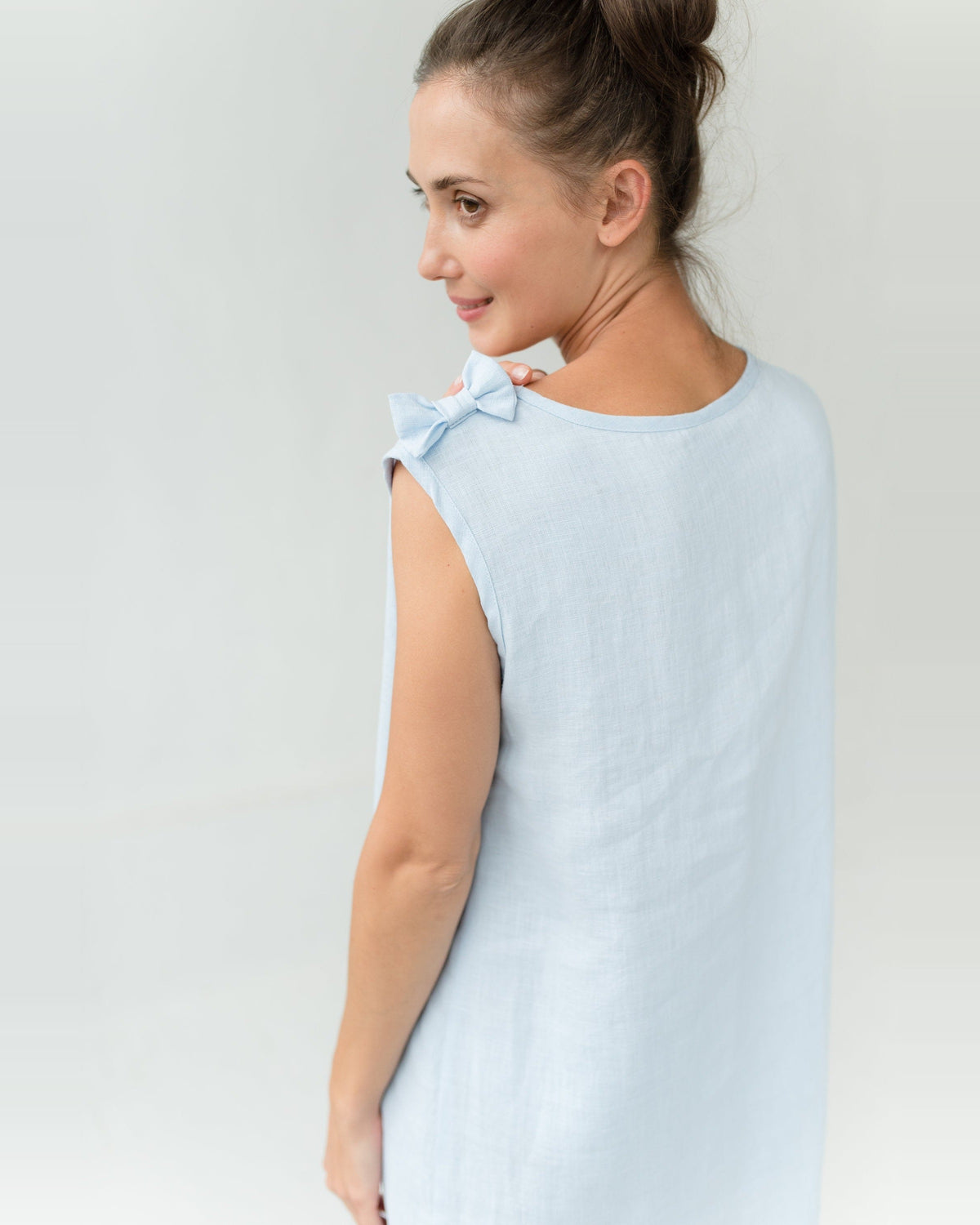 Femme en chemise de nuit en lin bleu clair à large décolleté plongeant en V et petit nœud sur l’épaule, debout les bras croisés.