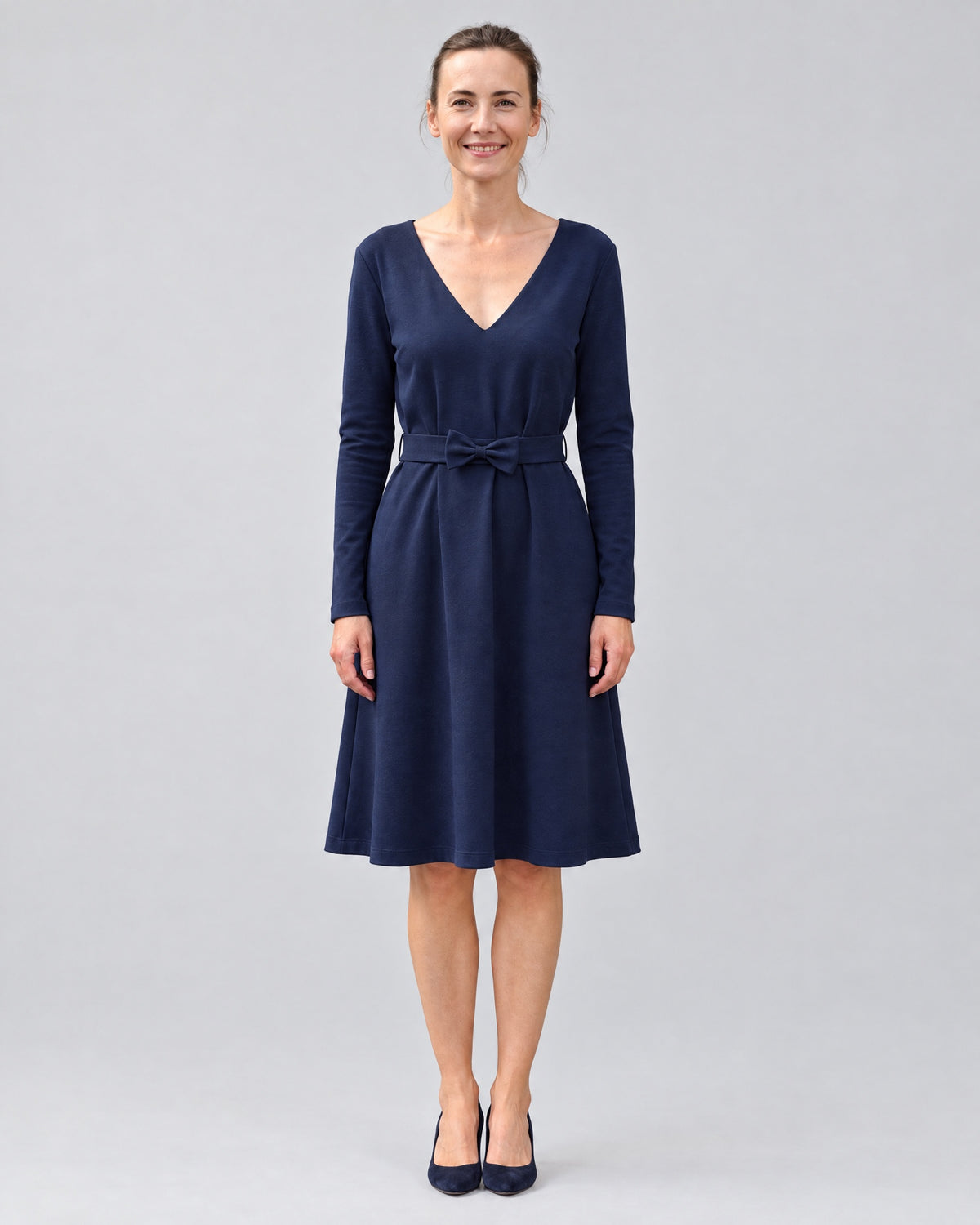 A woman wearing a navy blue long-sleeve V-neck Punto Milano dress with a tie waist, standing in a studio, front view, full length. #Color_Deep blue