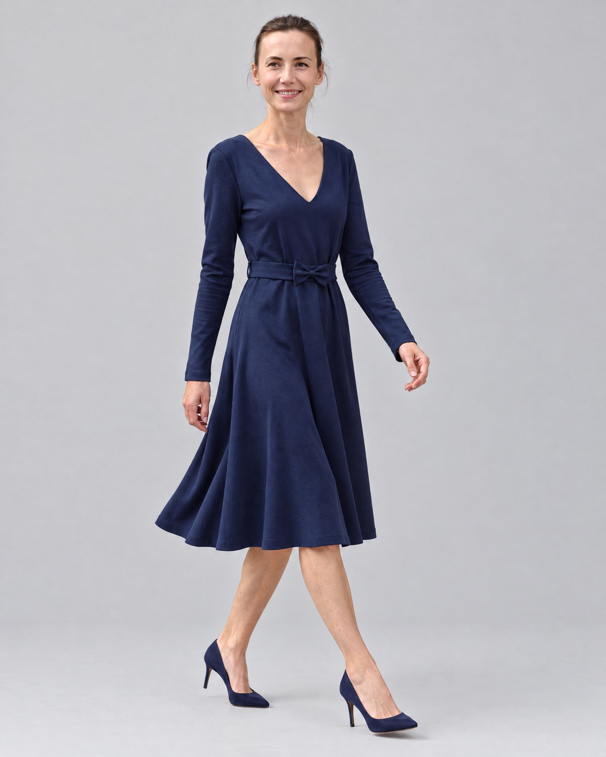 A woman walking in a navy blue long-sleeve Punto Milano dress, side view showing the A-line skirt in motion. #Color_Deep blue