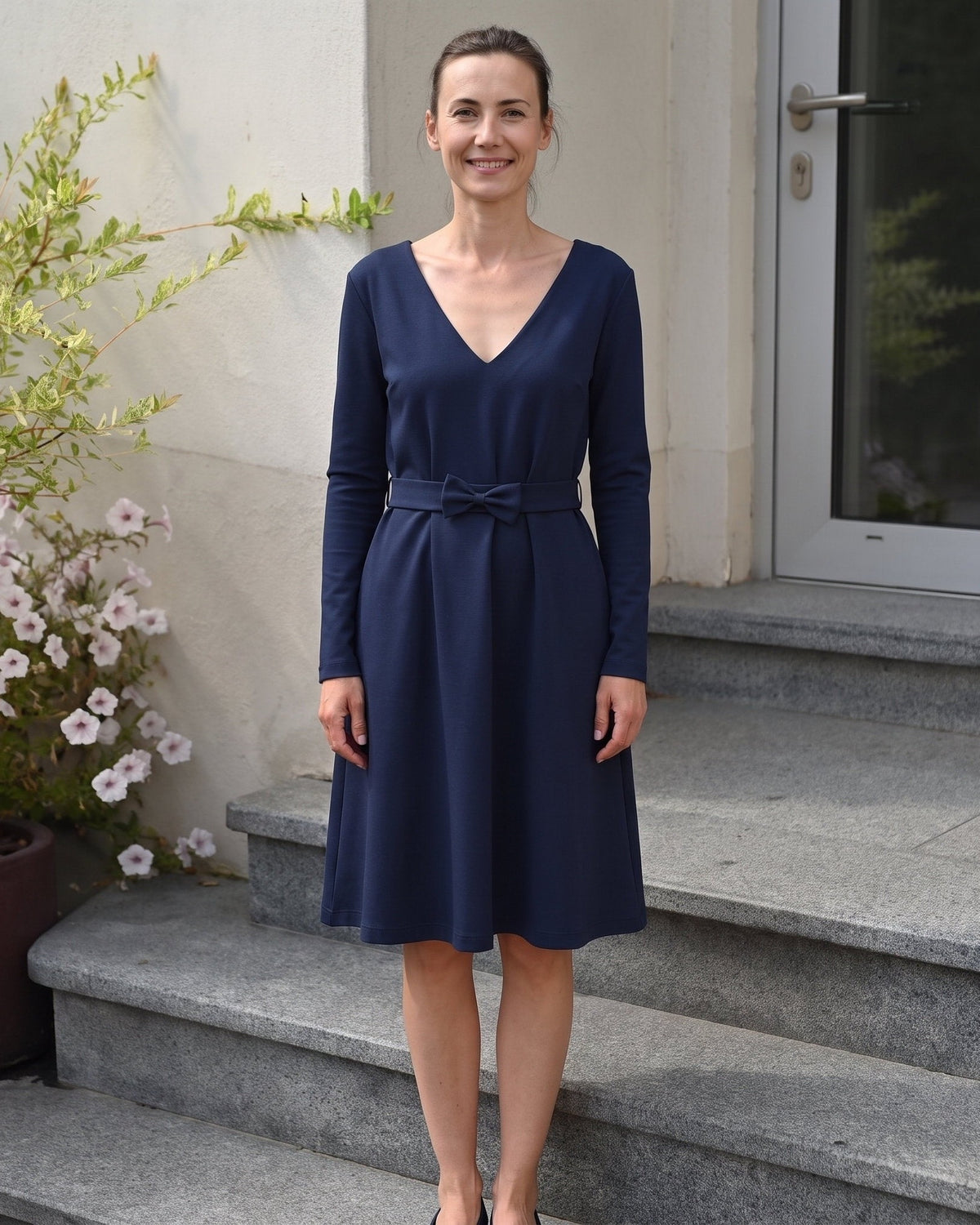 A woman wearing a navy blue long-sleeve V-neck dress with a tie waist, standing on stone steps in front of a building entrance. #Color_Deep blue
