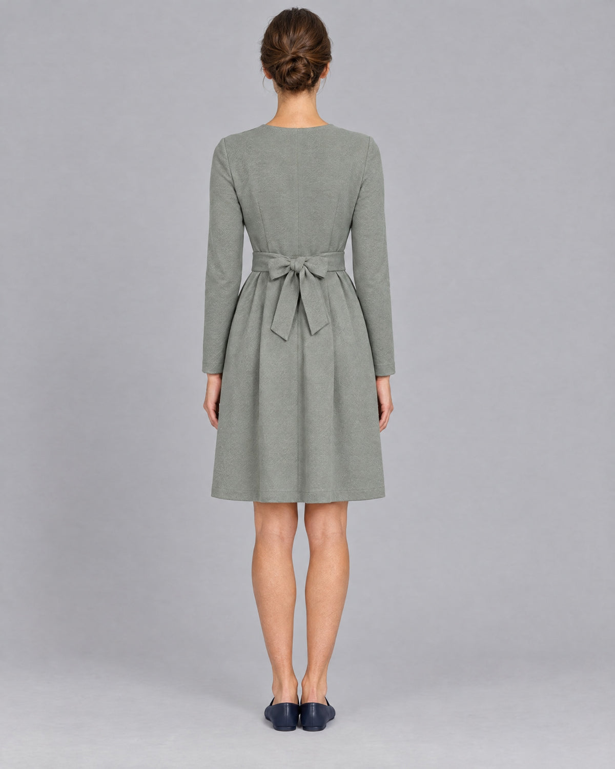 A woman wearing a sage gray-green long-sleeve V-neck Punto Milano dress with a tie belt bow, knee-length A-line skirt, back view, full length. #Color_Gray green