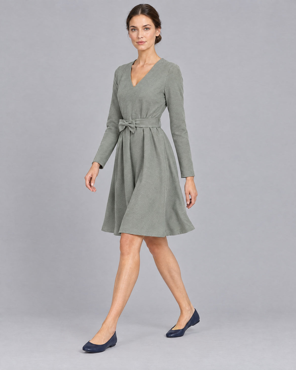 A woman walking in a sage gray-green long-sleeve Punto Milano dress, knee-length A-line skirt in motion, side view. #Color_Gray green