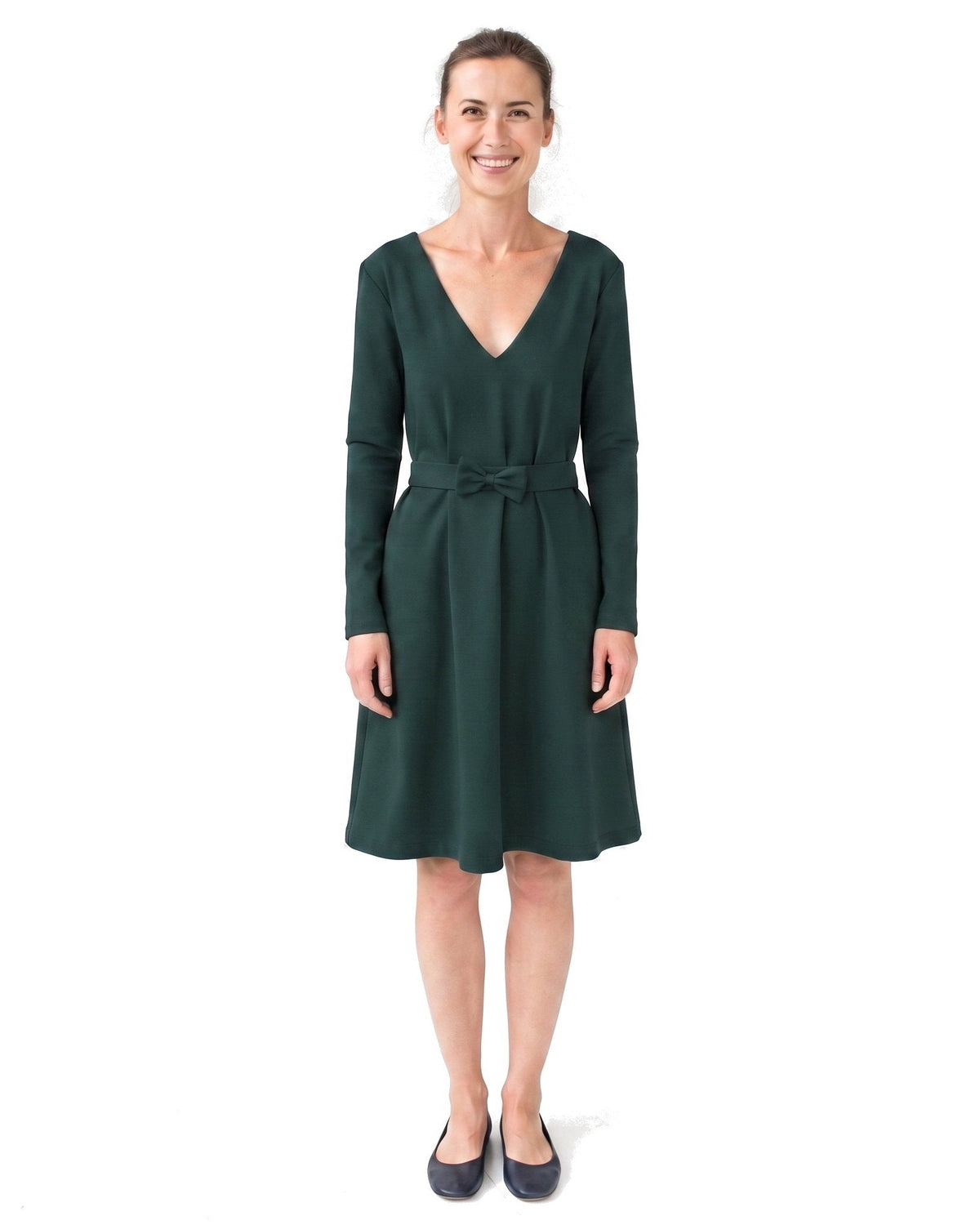 A woman modeling a dark green long-sleeve V-neck dress with a matching waist tie, paired with dark navy flats, against a plain white background. #Color_Green