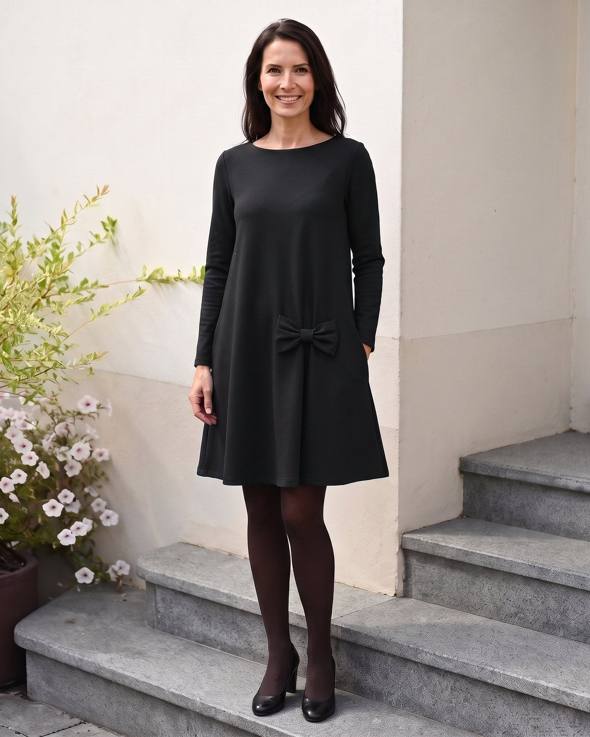 A woman stands on stone steps wearing a black long-sleeve knee-length dress with a bow detail at the front. #Color_Black