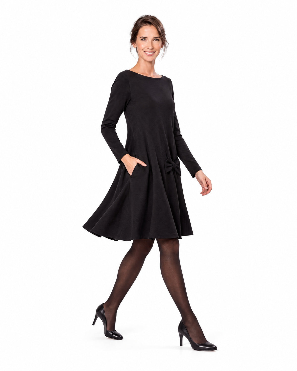 A woman modeling a black long-sleeve A-line dress with a boat neckline and bow-detail pocket, shown in profile while walking. #Color_Black
