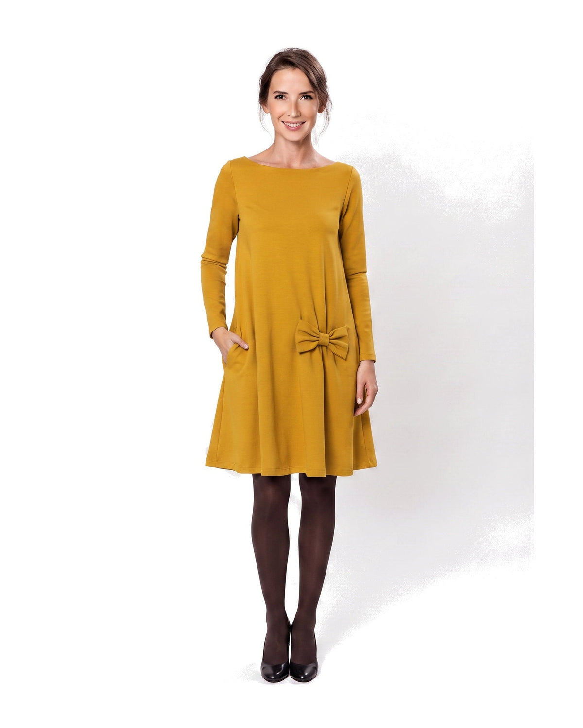 A woman modeling a mustard yellow long-sleeve A-line dress with a boat neckline, side pockets, and a bow detail at the waist. #Color_Yellow