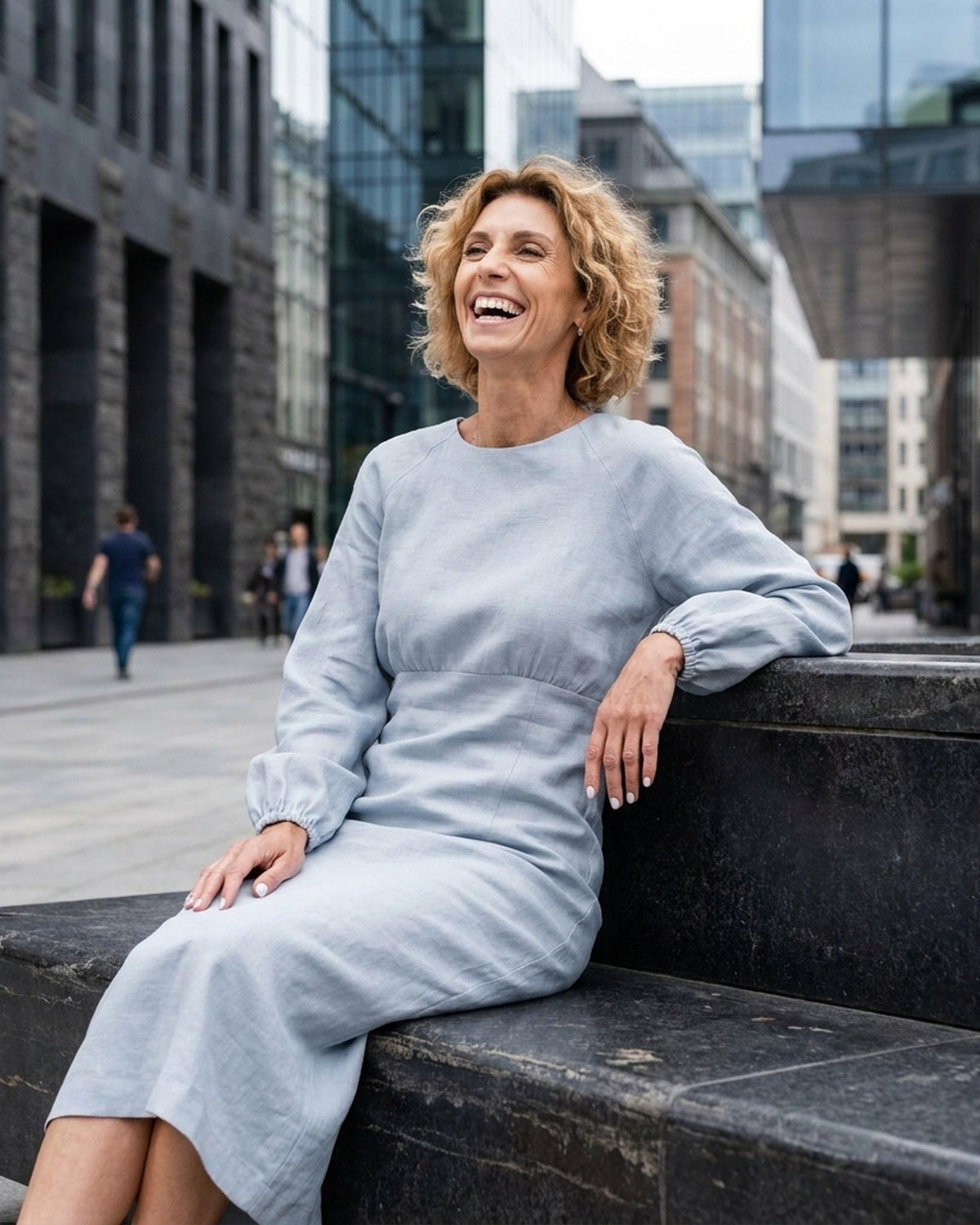 A woman with short blonde hair sits smiling, wearing a light blue long-sleeve midi dress with a gathered waist and elastic cuffs. #Color_Light blue
