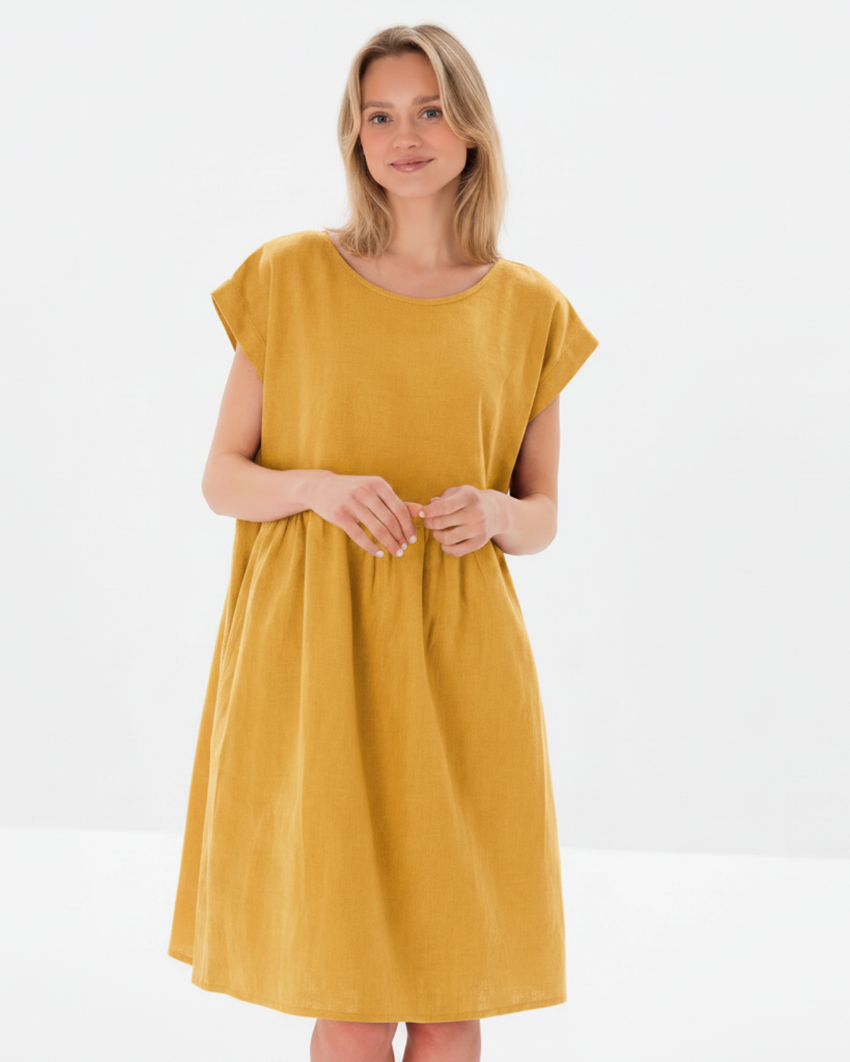 A woman modeling a mustard yellow linen dress with a relaxed, loose fit, short cap sleeves, and a round neckline against a clean white studio background.