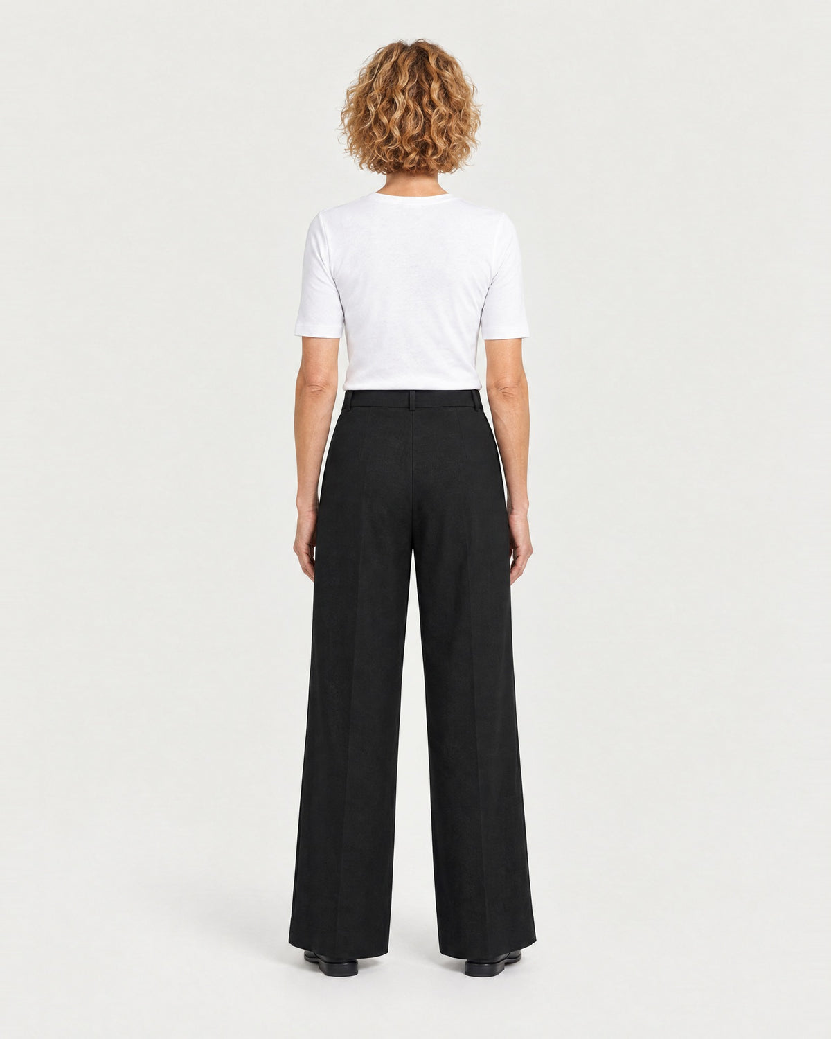 A woman stands with her back to the camera against a light gray studio background, wearing a white short-sleeve crewneck top and black wide-leg trousers with a plain back and belt loops. #Color_Black