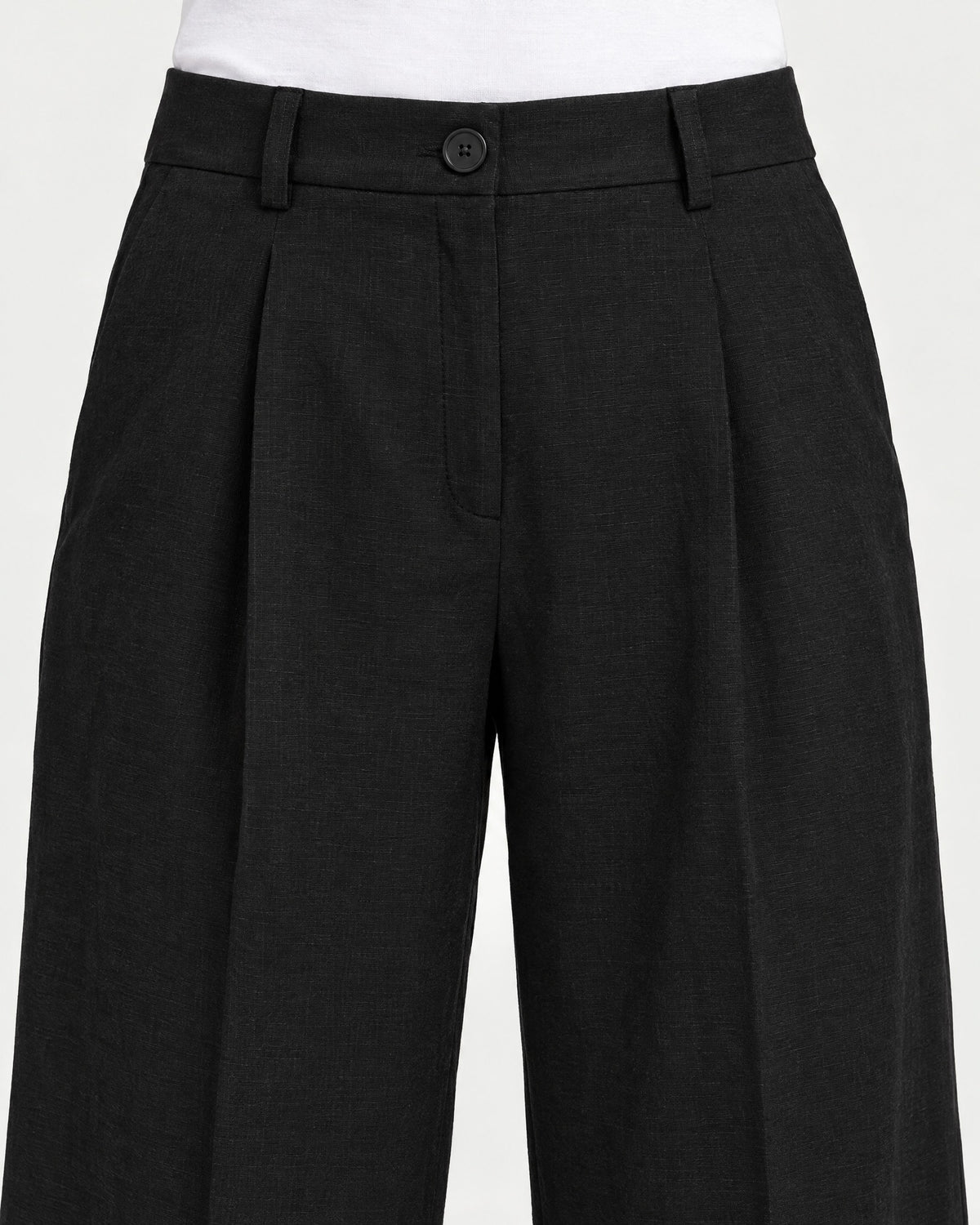 Close-up of black wide-leg linen trousers showing the waistband, front pleats and button closure. #Color_Black