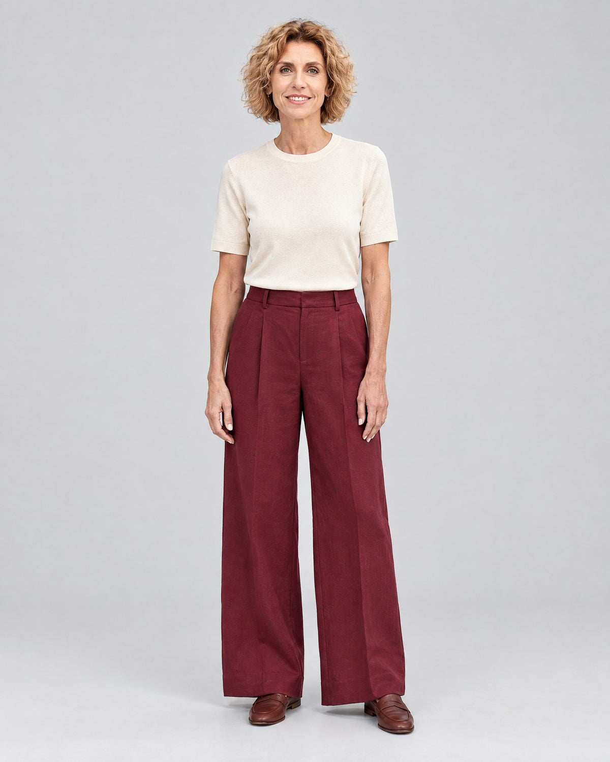 A woman stands facing the camera against a light gray studio background, wearing a cream short-sleeve top, burgundy wide-leg trousers and brown loafers. #Color_Burgundy