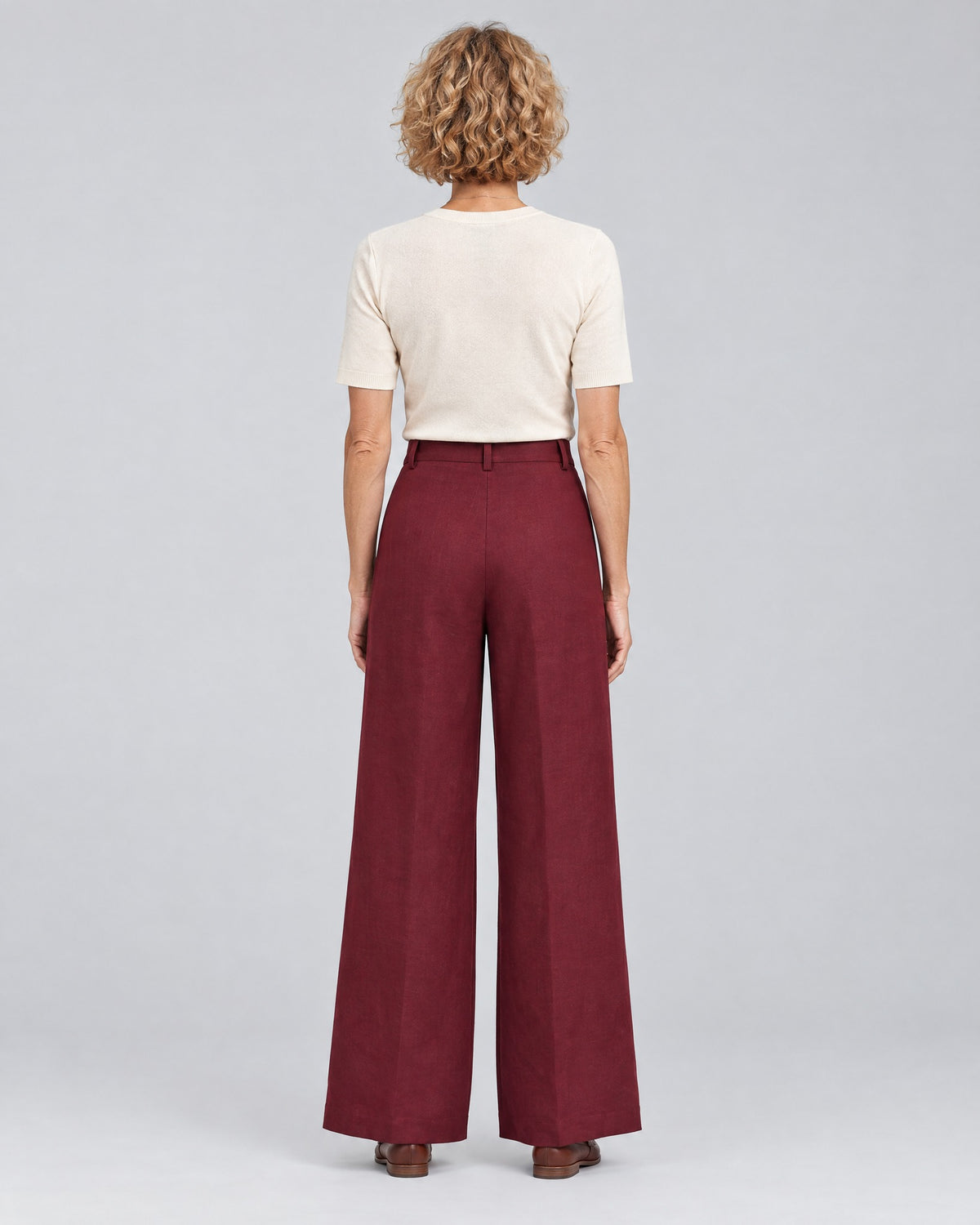 A woman stands with her back to the camera against a light gray studio background, wearing a cream short-sleeve top and burgundy wide-leg trousers with a plain back and belt loops. #Color_Burgundy