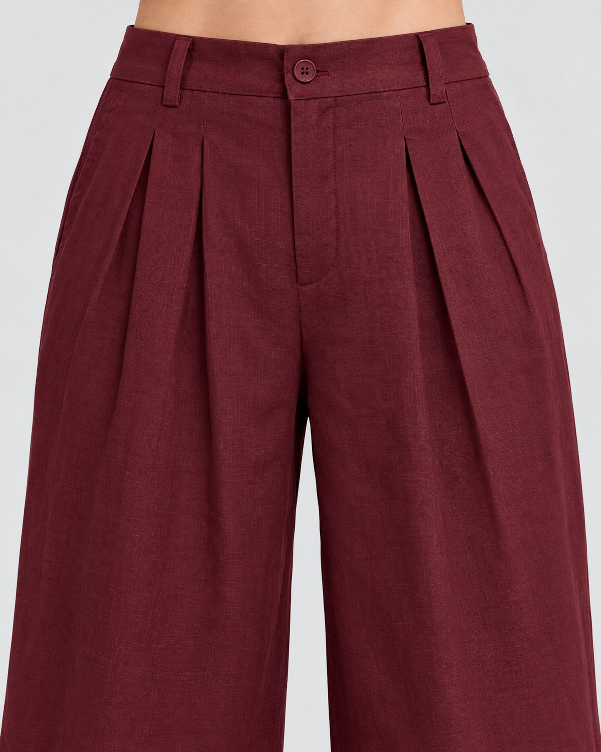 Close-up of burgundy wide-leg linen trousers showing the waistband, front pleats and button closure. #Color_Burgundy