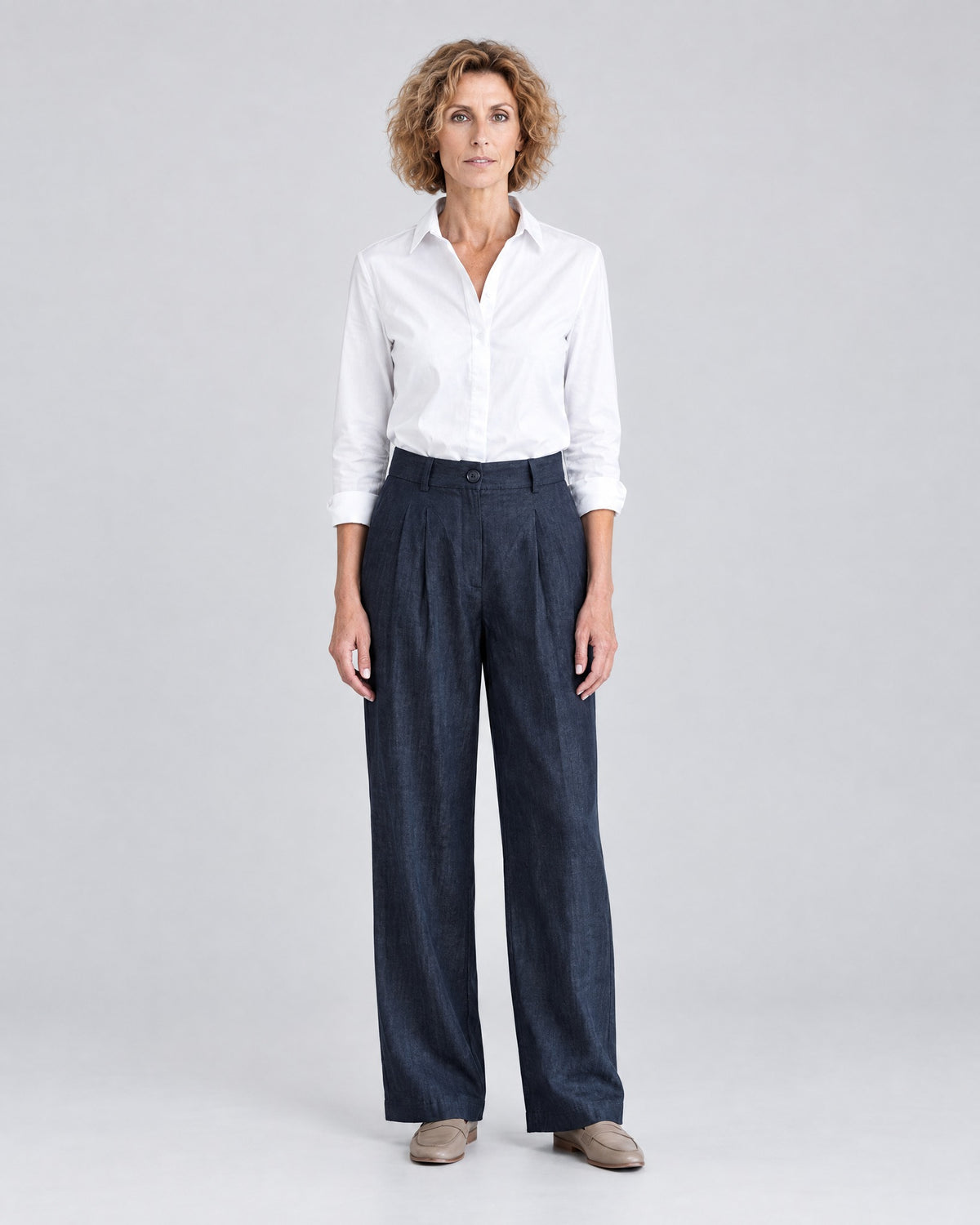 A woman stands facing the camera against a light gray studio background, wearing a white button-up shirt, deep navy blue wide-leg trousers and tan loafers. #Color_Deep blue