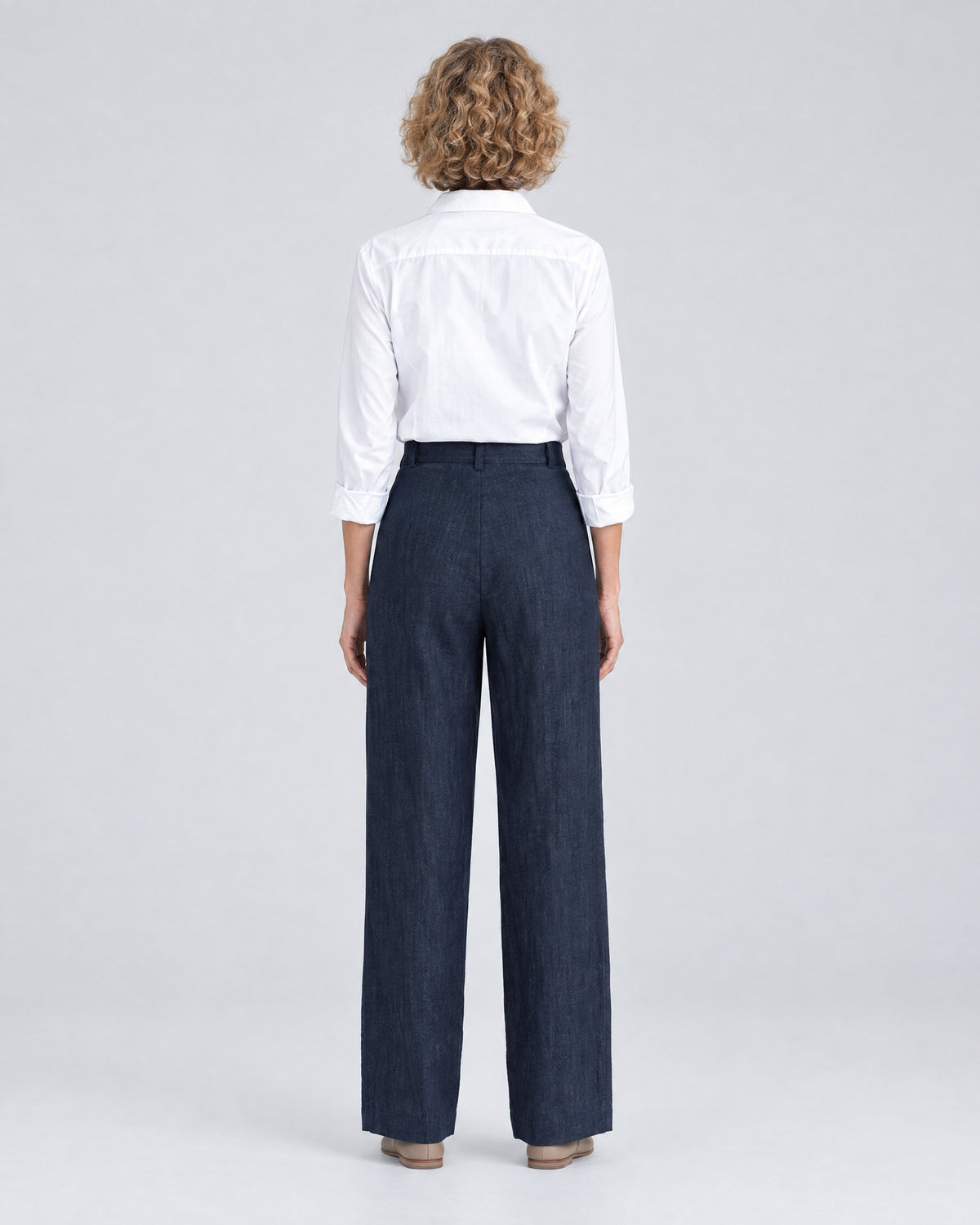 A woman stands with her back to the camera against a light gray studio background, wearing a white button-up shirt and deep navy blue wide-leg trousers with a plain back and belt loops. #Color_Deep blue
