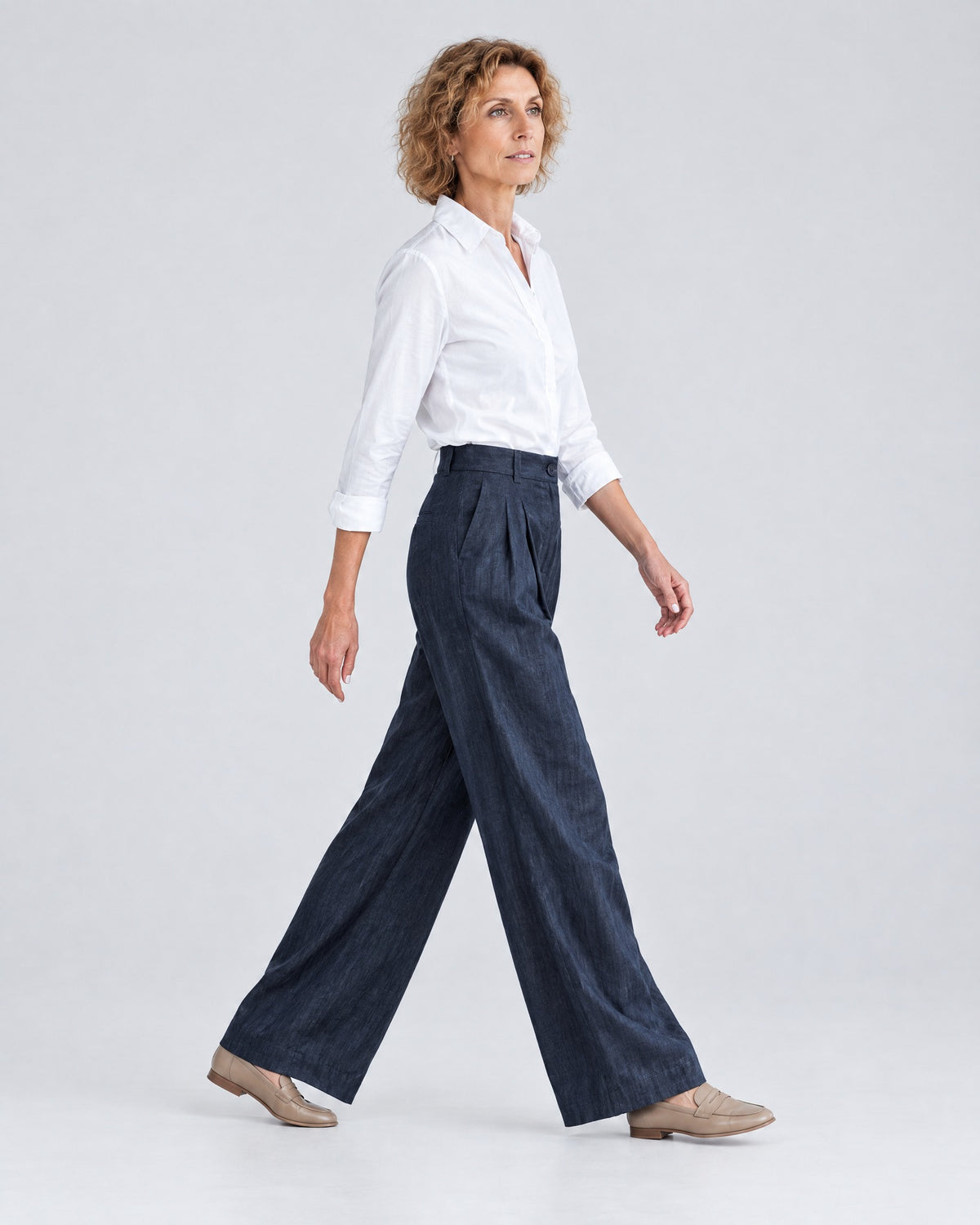 A woman walks mid-stride in profile against a light gray studio background, wearing a white button-up shirt, deep navy blue wide-leg trousers and tan loafers. #Color_Deep blue