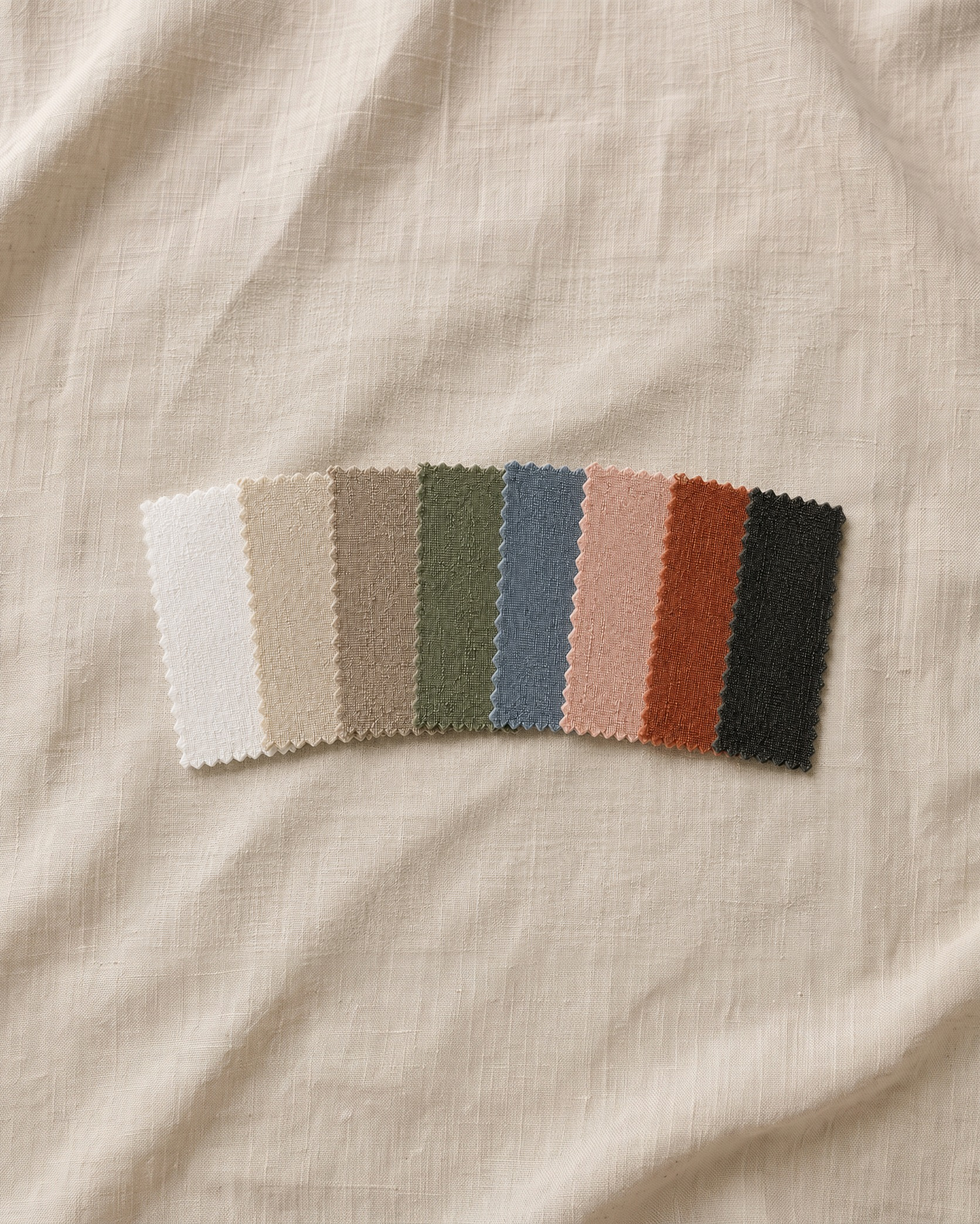 Huit échantillons de tissu texturé dans une palette chaude, neutre et terreuse, disposés en éventail sur un fond de lin beige. Les couleurs comprennent blanc cassé, crème, camel, vert sauge, bleu poudré, rose blush, orange terracotta et noir charbon.